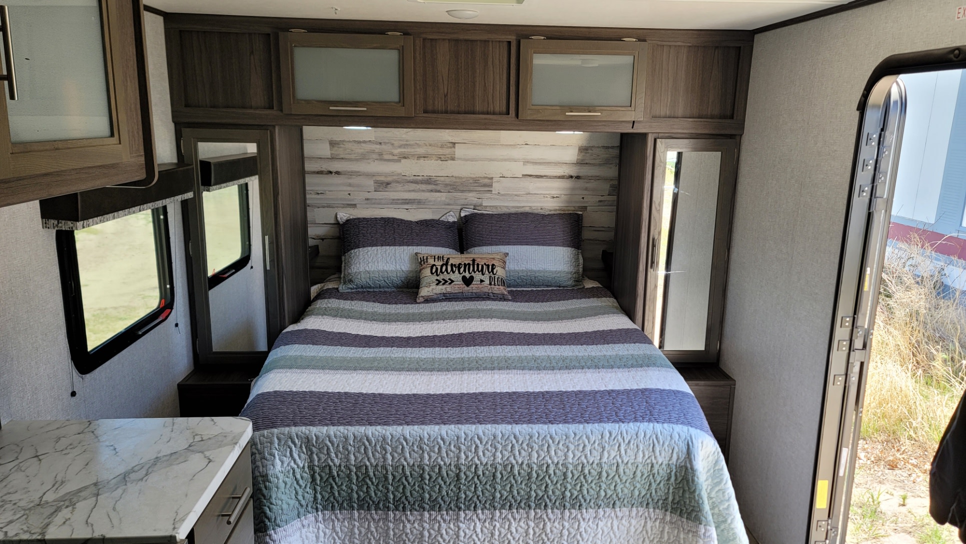 Cozy RV rental bedroom with striped bed, "adventure" pillow, dark wood cabinets, and light walls with windows.. Dutchmen Kodiak Ultra Lite 2020