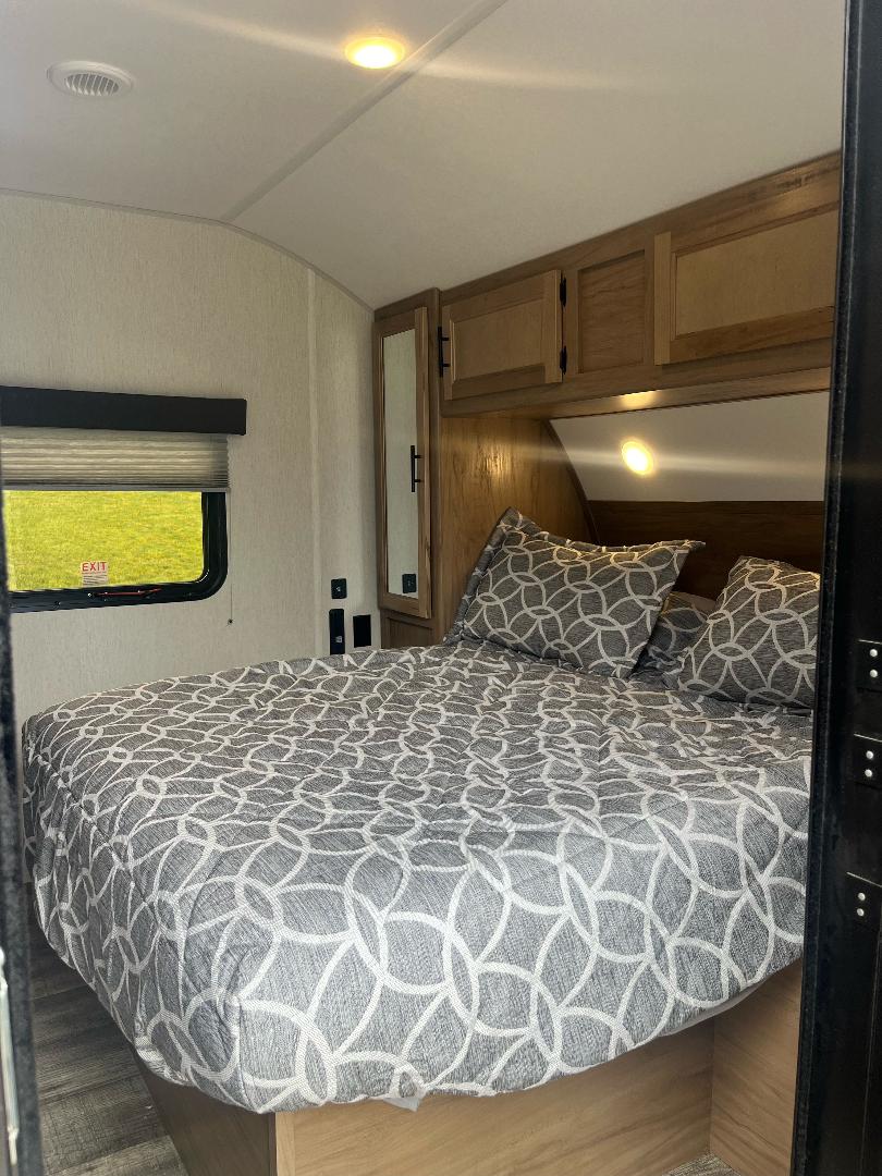A well-appointed RV rental bedroom with a large bed featuring gray patterned bedding, overhead cabinets, and a window revealing green outdoors.. Forest River Cherokee Grey Wolf 2023