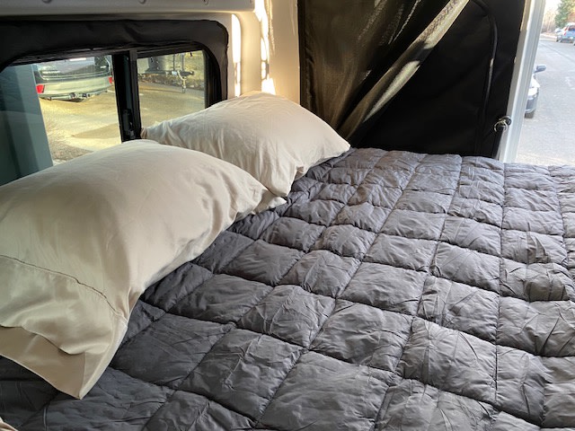 Inside an RV rental: a comfortable bed with light pillows & a dark grey quilted comforter, perfect for travel.. Winnebago Solis 2021