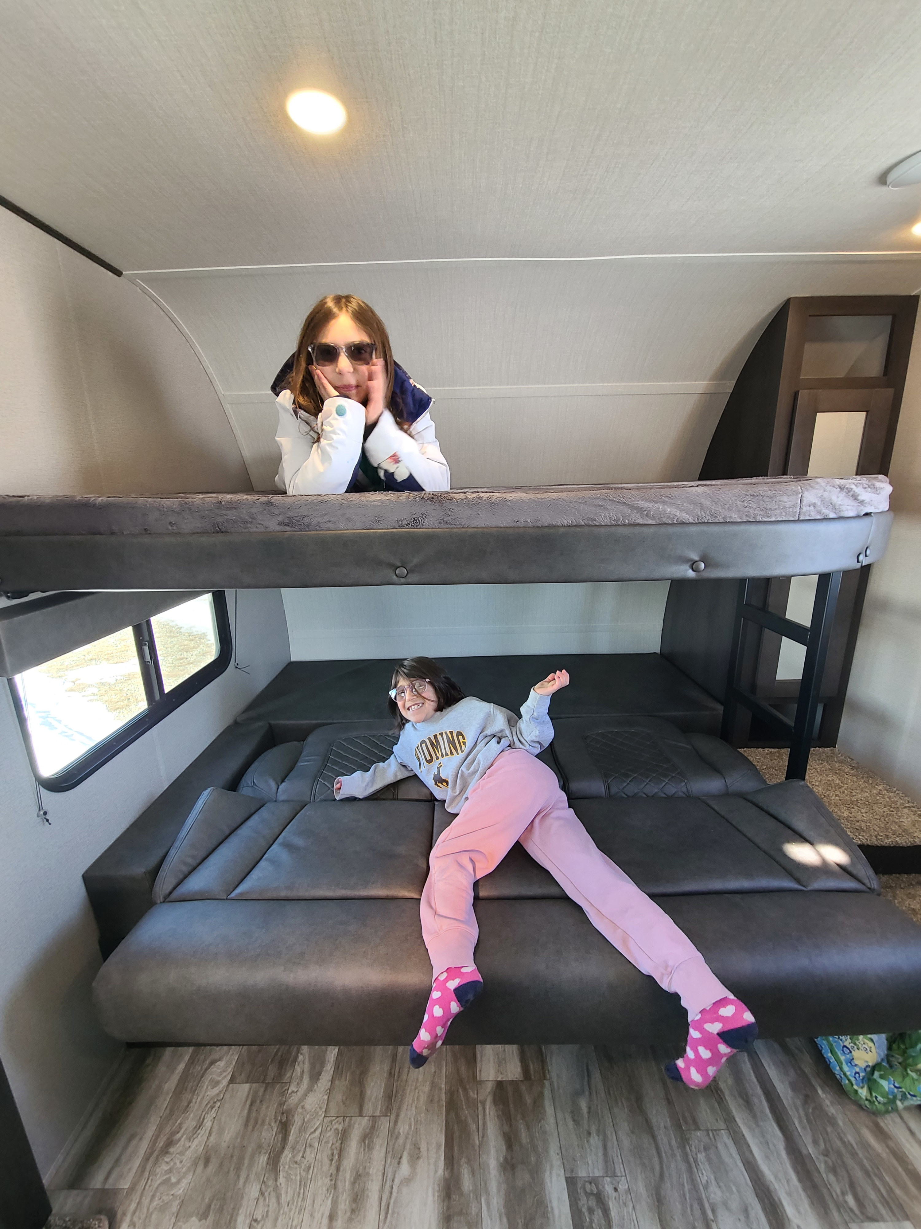 Inside an RV rental, two girls pose. One on the upper bunk with shades, the other on the lower bed in a Wyoming shirt.. Grand Design Transcend Xplor 2022