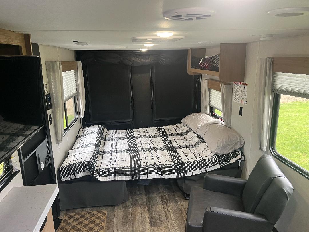 RV rental interior features a cozy bed with plaid comforter, black wall panels, and an armchair, offering a comfortable stay.. Forest River Cherokee Grey Wolf 2023
