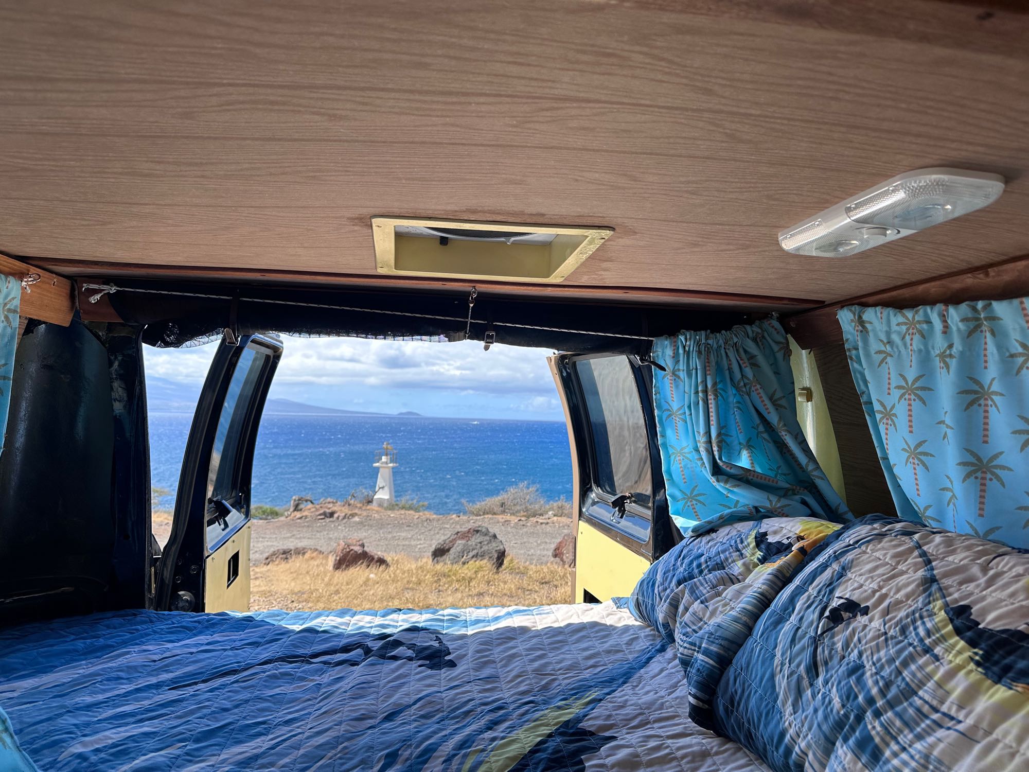 RV rental: Inside a camper van, a cozy bed faces open doors revealing a distant lighthouse and expansive ocean.. Dodge 1500 1999