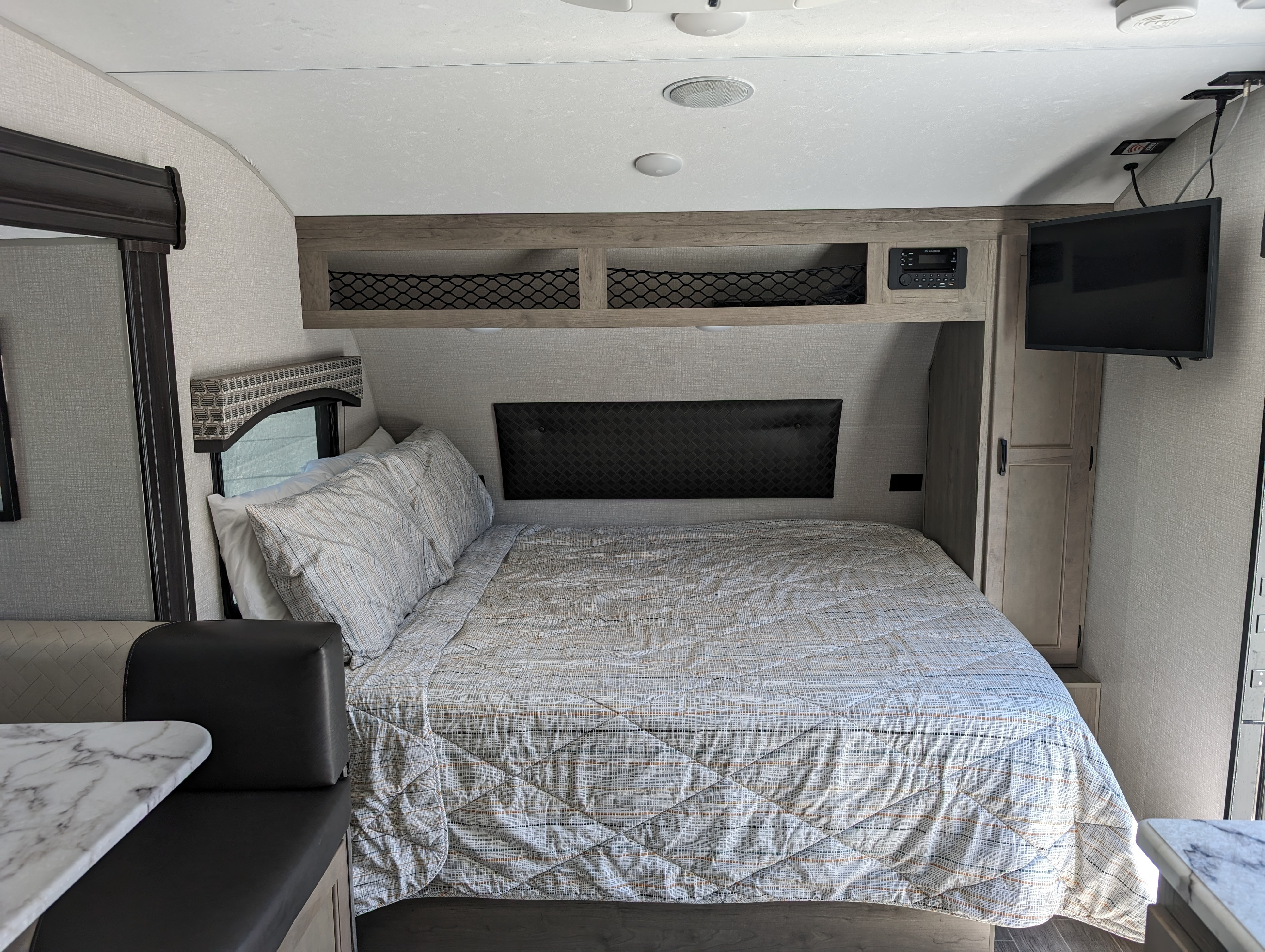 A compact bedroom in an RV rental, featuring a comfortable bed, wall-mounted TV, overhead storage, and a dinette.. Sonic Lite 2021