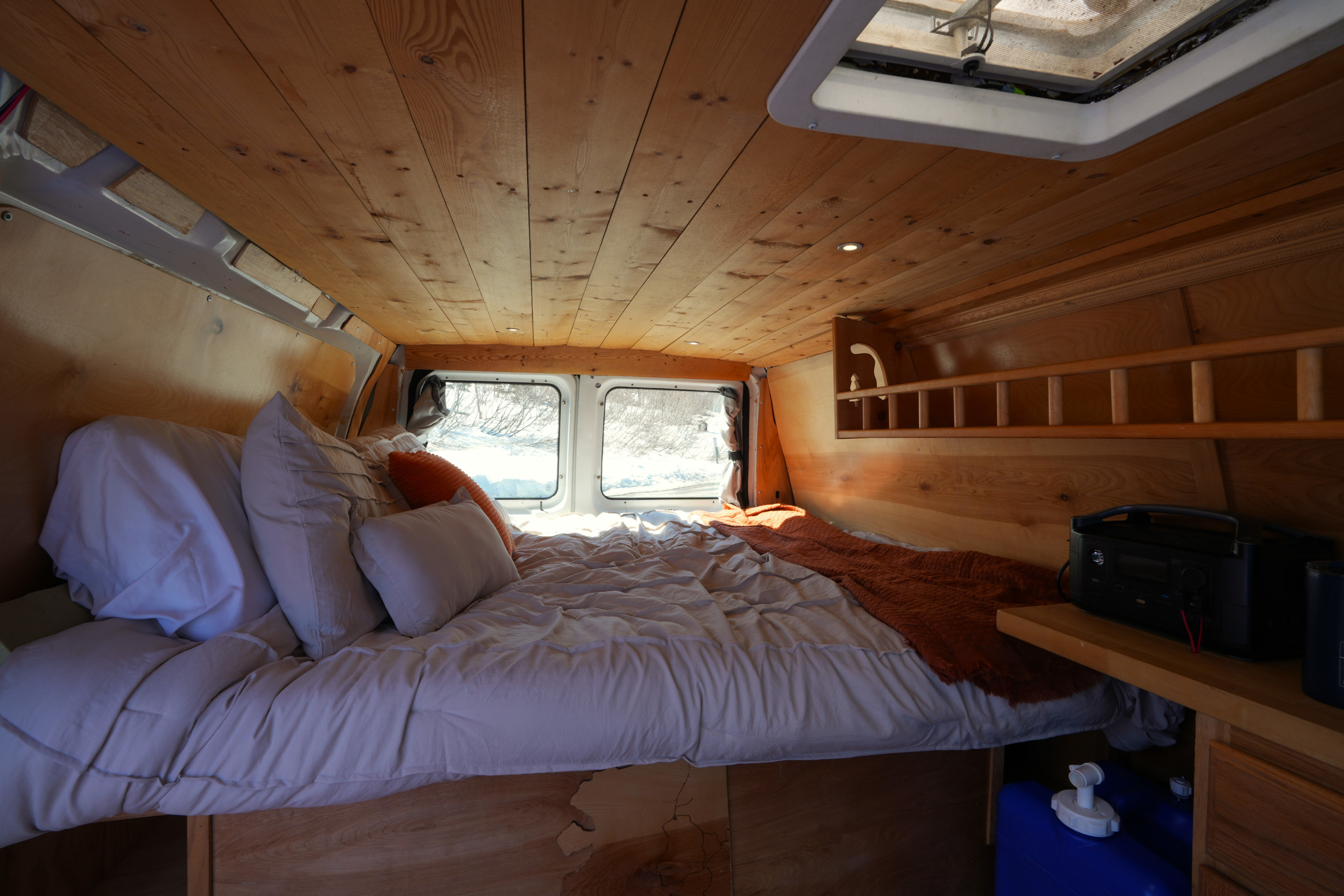Inside a cozy RV rental: a wooden-paneled camper van bedroom with a comfortable bed, pillows, and snowy window views.. Ford E250 Camper Van 2001