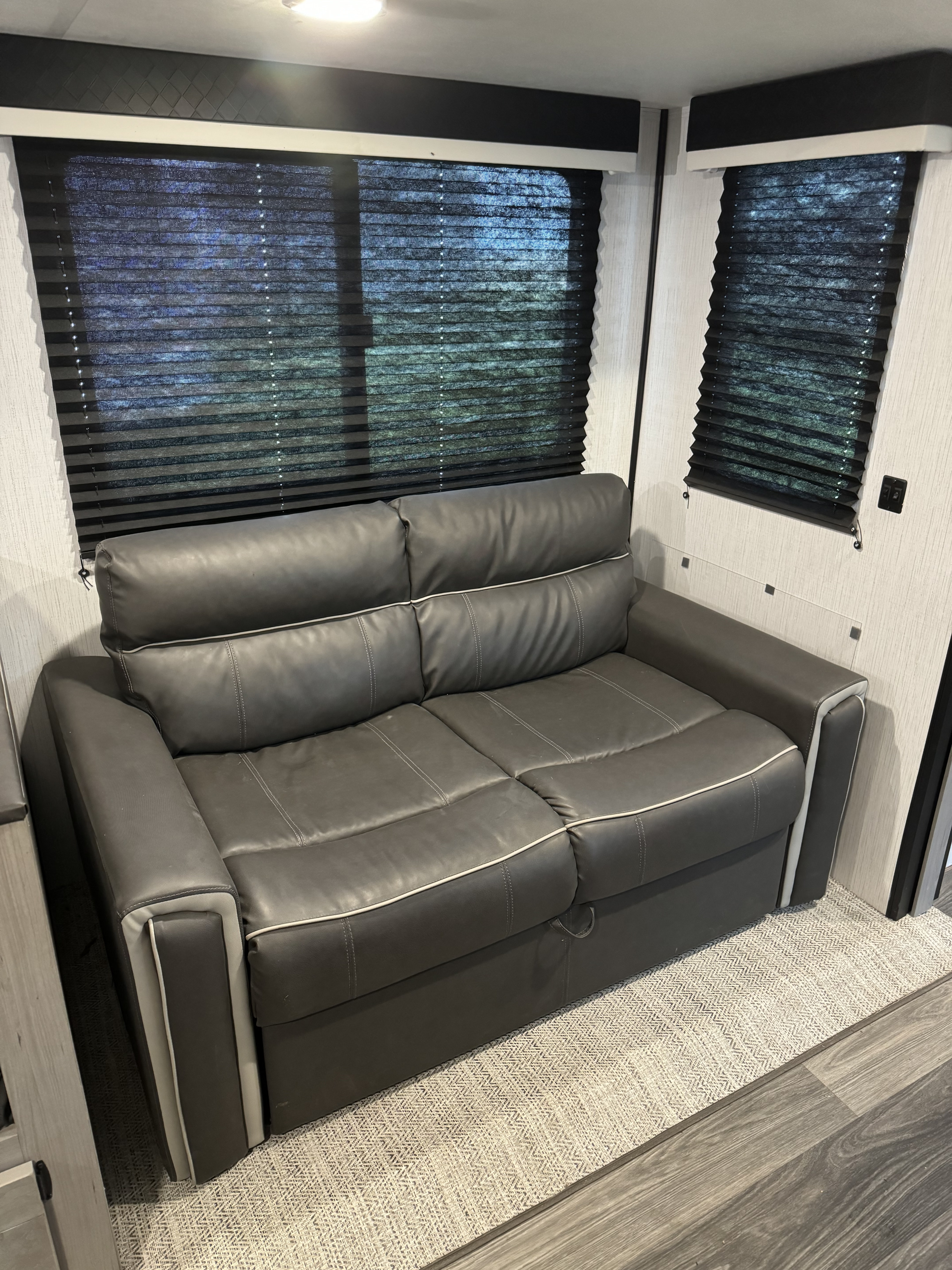 Comfortable two-seater gray sofa in an RV rental, featuring pleated blinds and wood flooring. Ideal for travel.. Heartland RVs Mallard 2022