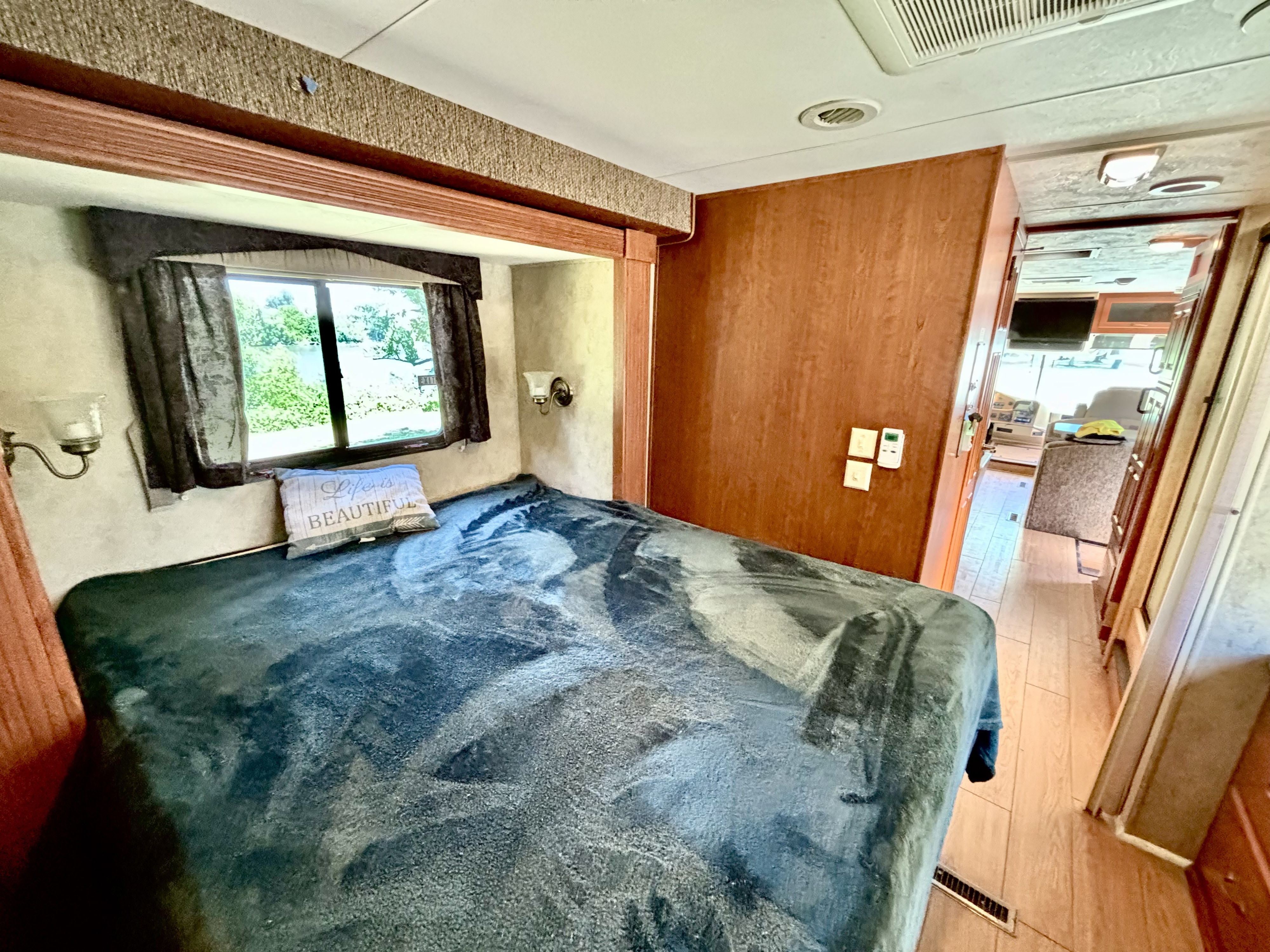 RV rental bedroom with a large bed, window showing greenery, wood walls, and hallway to living space.. Damon Daybreak 2009