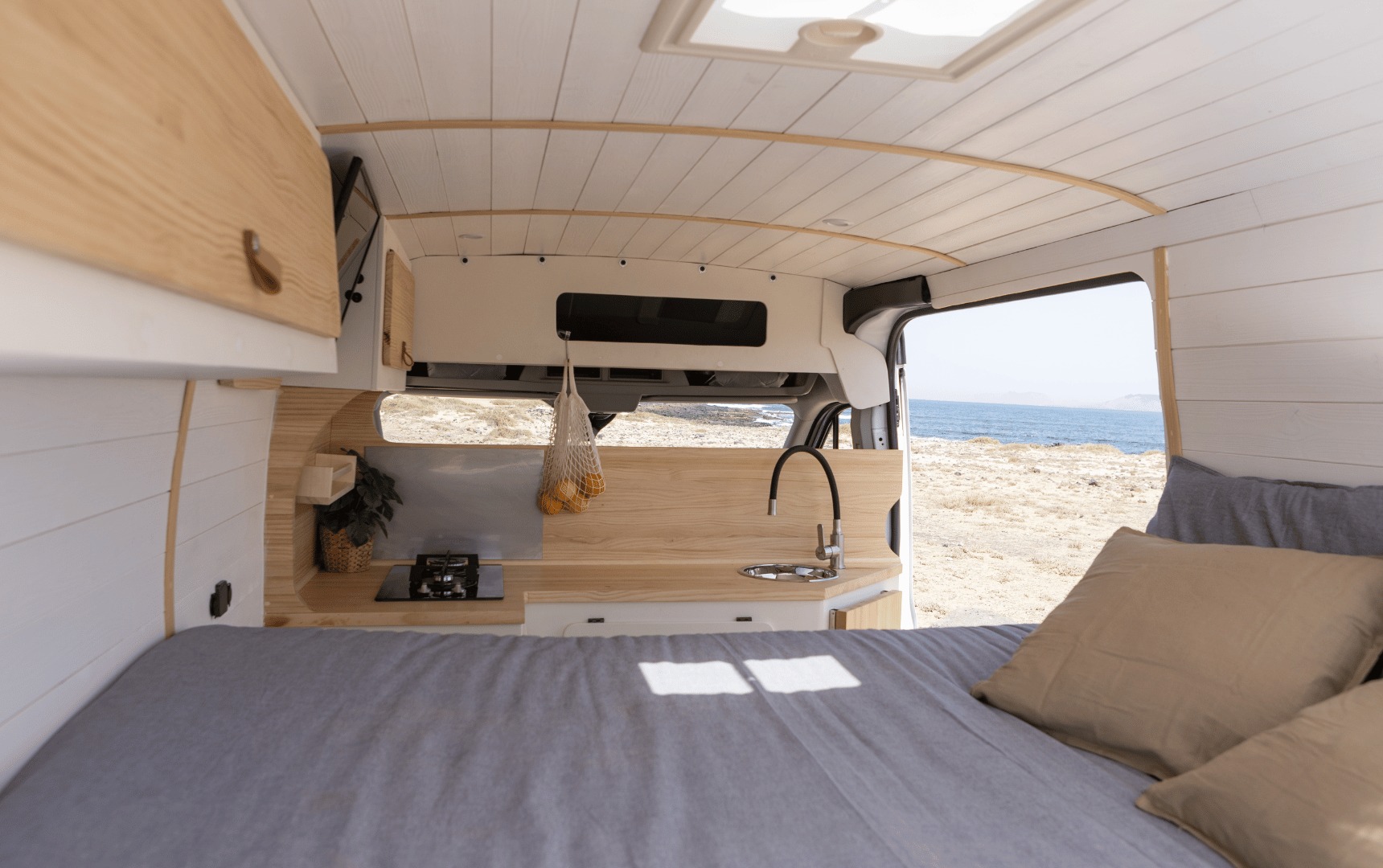 Bright RV rental interior: wood and white finishes, a comfy bed, modern kitchen, and an ocean view.. Ford Transit 2023
