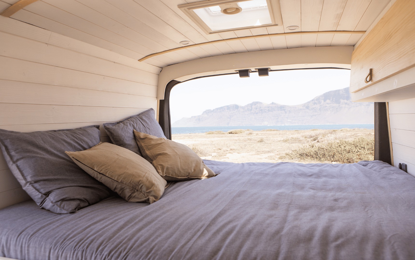 Cozy **RV rental** bedroom with a comfy bed & open back doors revealing panoramic ocean & mountain views. Ideal for van life adventures.. Ford Transit 2023