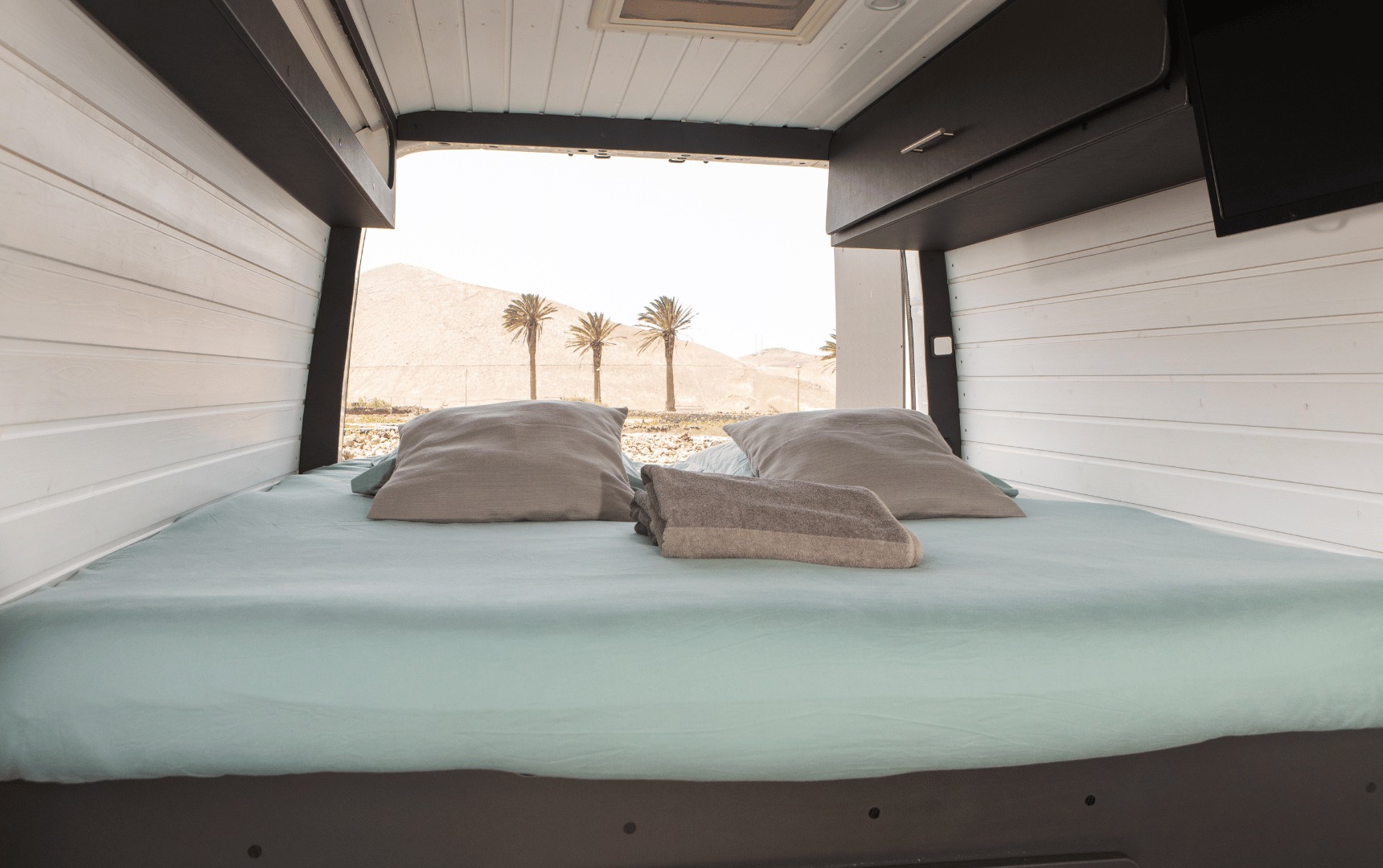 RV rental interior: A cozy bed with blue sheets & grey pillows overlooks desert mountains & palm trees through open rear doors.. Volkswagen Crafter 2019