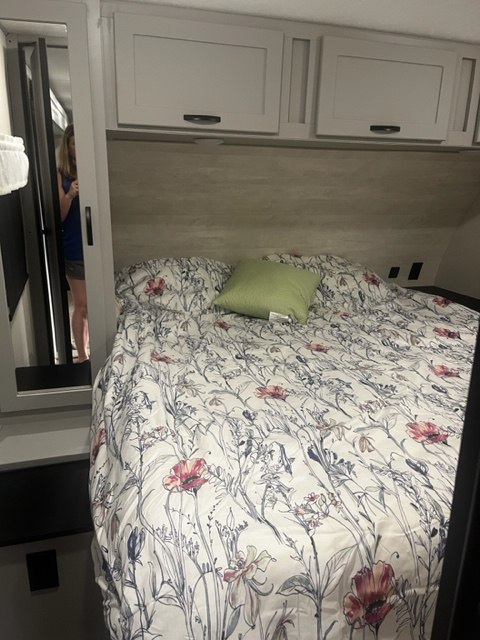 Cozy RV rental bedroom with floral bedding, overhead cabinets, and a mirror reflecting a person. Perfect for travel.. Forest River Vibe 2021