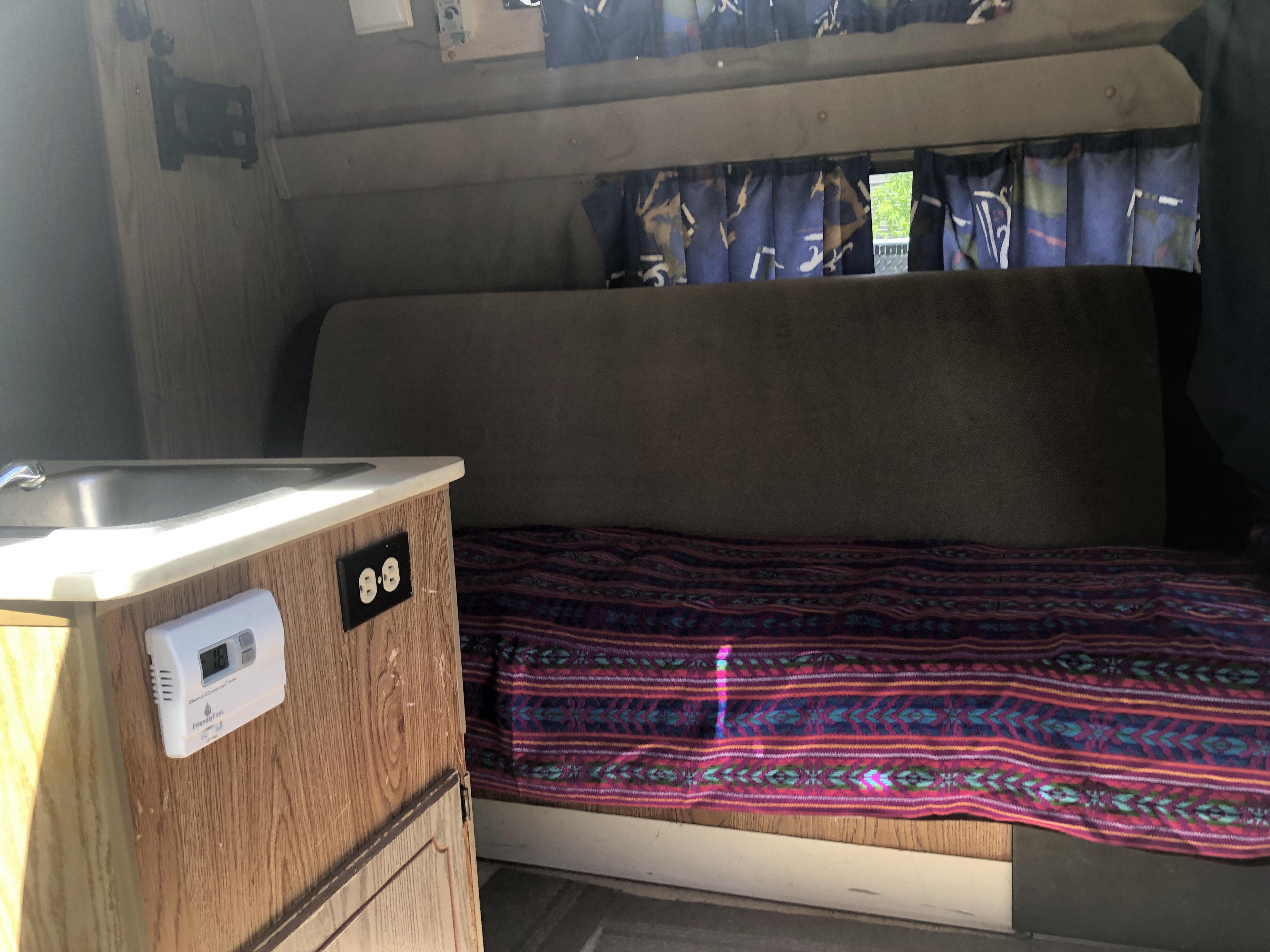 Discover this RV rental's interior: small sink, thermostat, vibrant bedding, and blue curtains. Ready for adventure!. Ford Econoline 150 1989