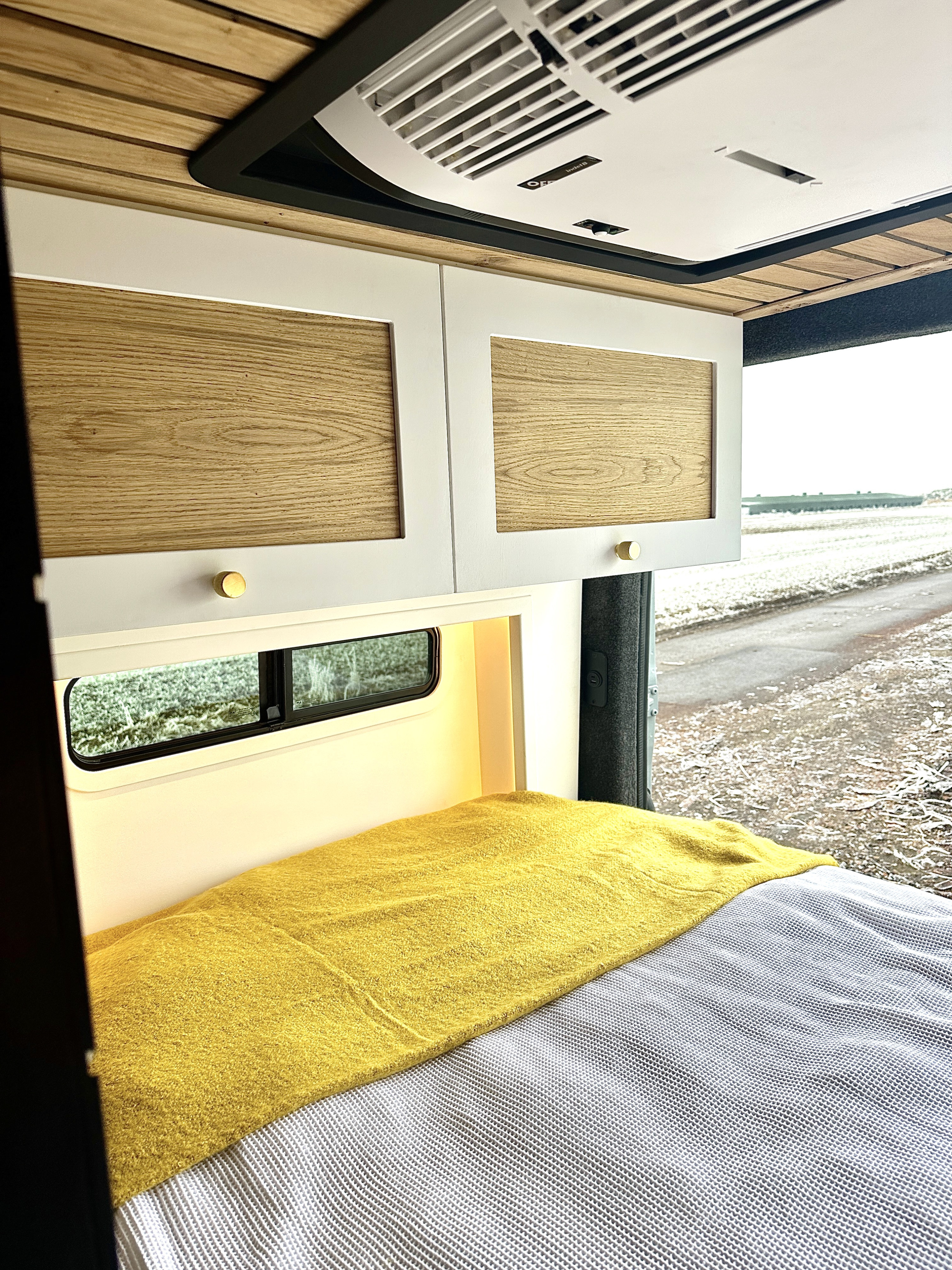Cozy RV rental interior featuring a bed with yellow throw, modern white cabinets, and overhead AC unit. Ideal for travel.. Mercedes Benz Sprinter 2023