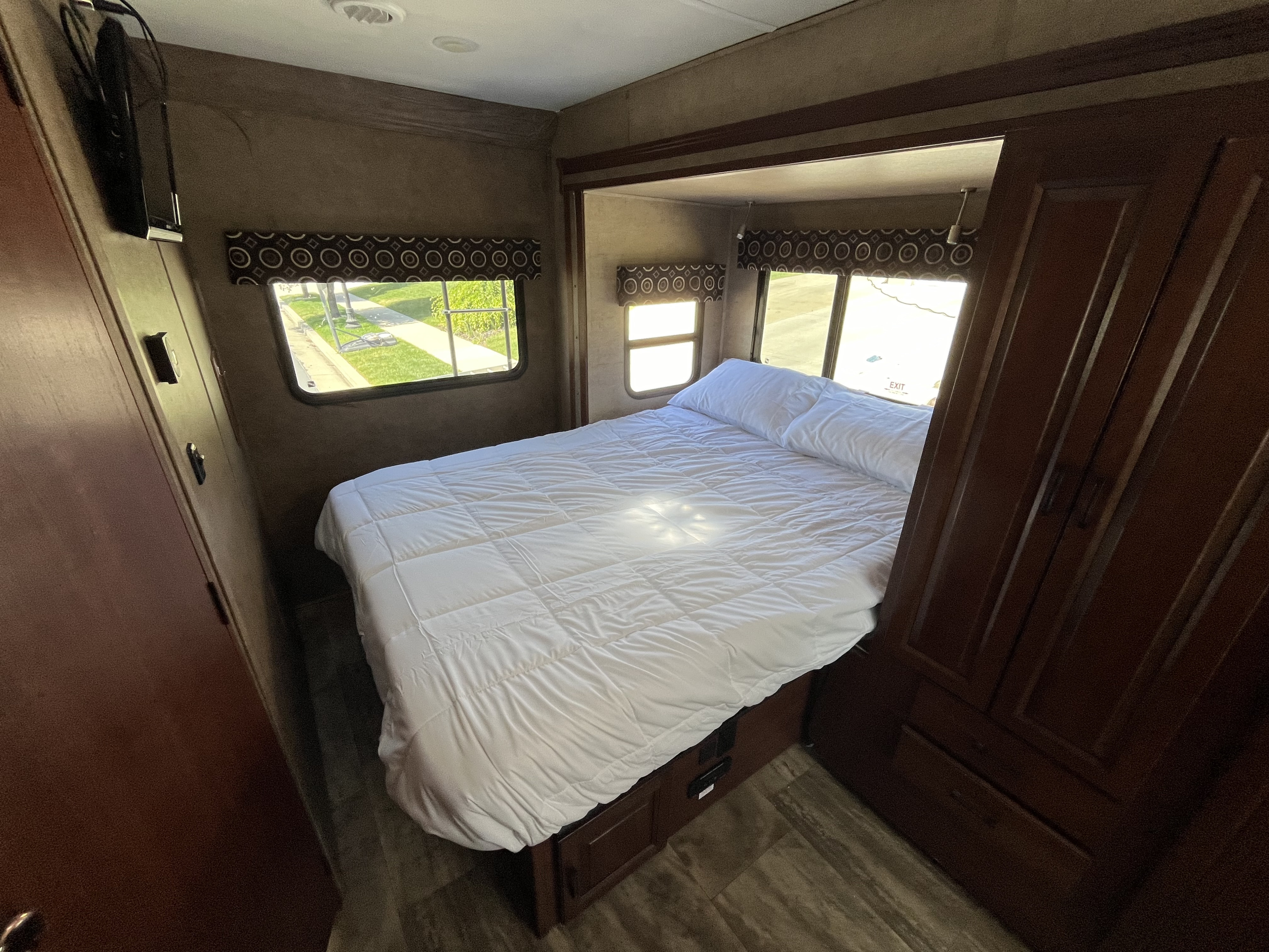Bright RV rental bedroom showcasing a queen bed with white bedding, side windows, a wall-mounted TV, and dark wood wardrobe.. Forest River Sunseeker MBS 2016