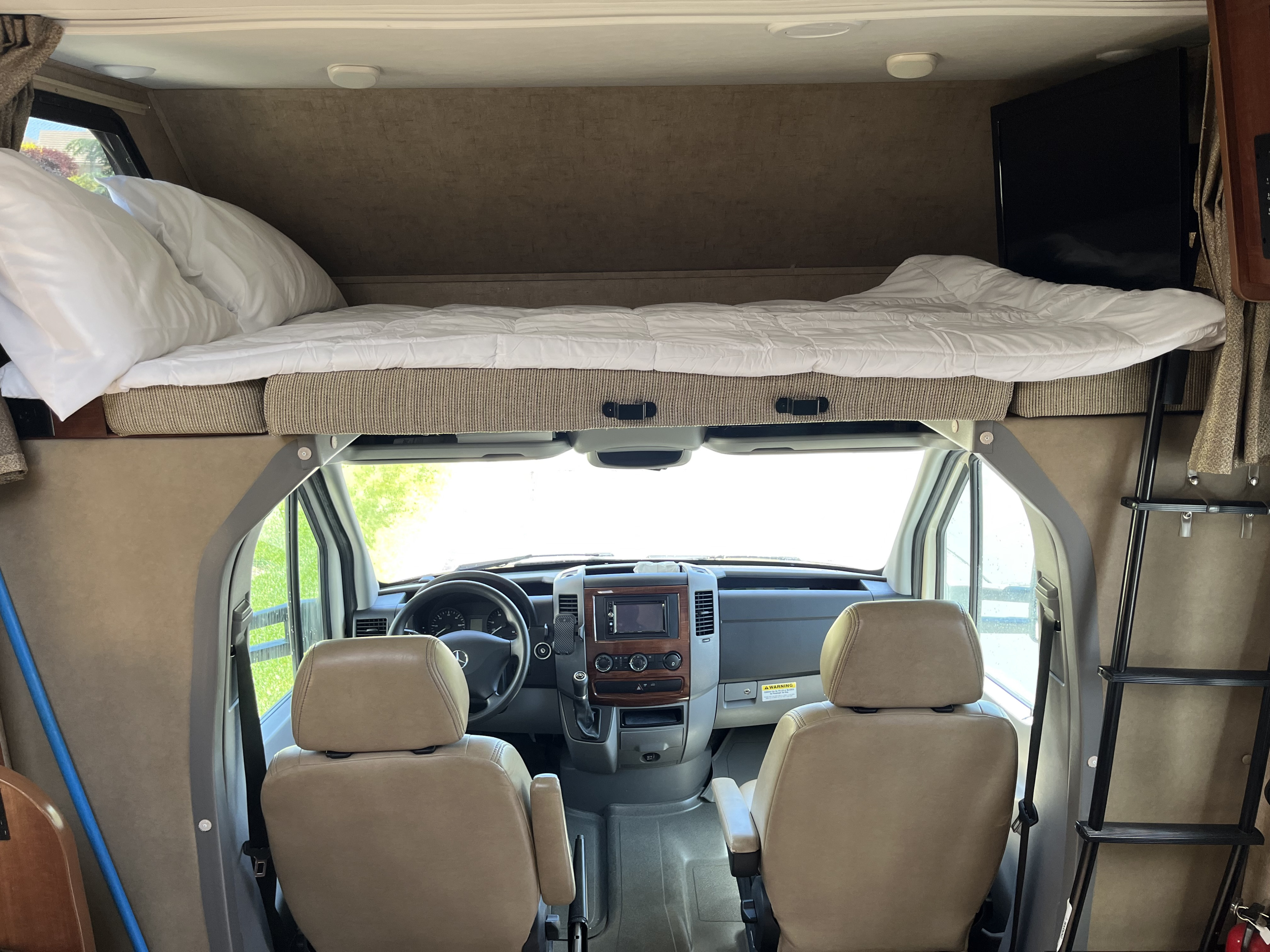 RV rental interior: cozy cab-over bed, driver's cabin with dashboard, and a TV on the right.. Forest River Sunseeker MBS 2016