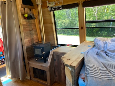 Experience a cozy RV rental interior: wood-paneled walls, a compact wood stove, and a comfortable bed by the window.. GMC Savana 3500 2011
