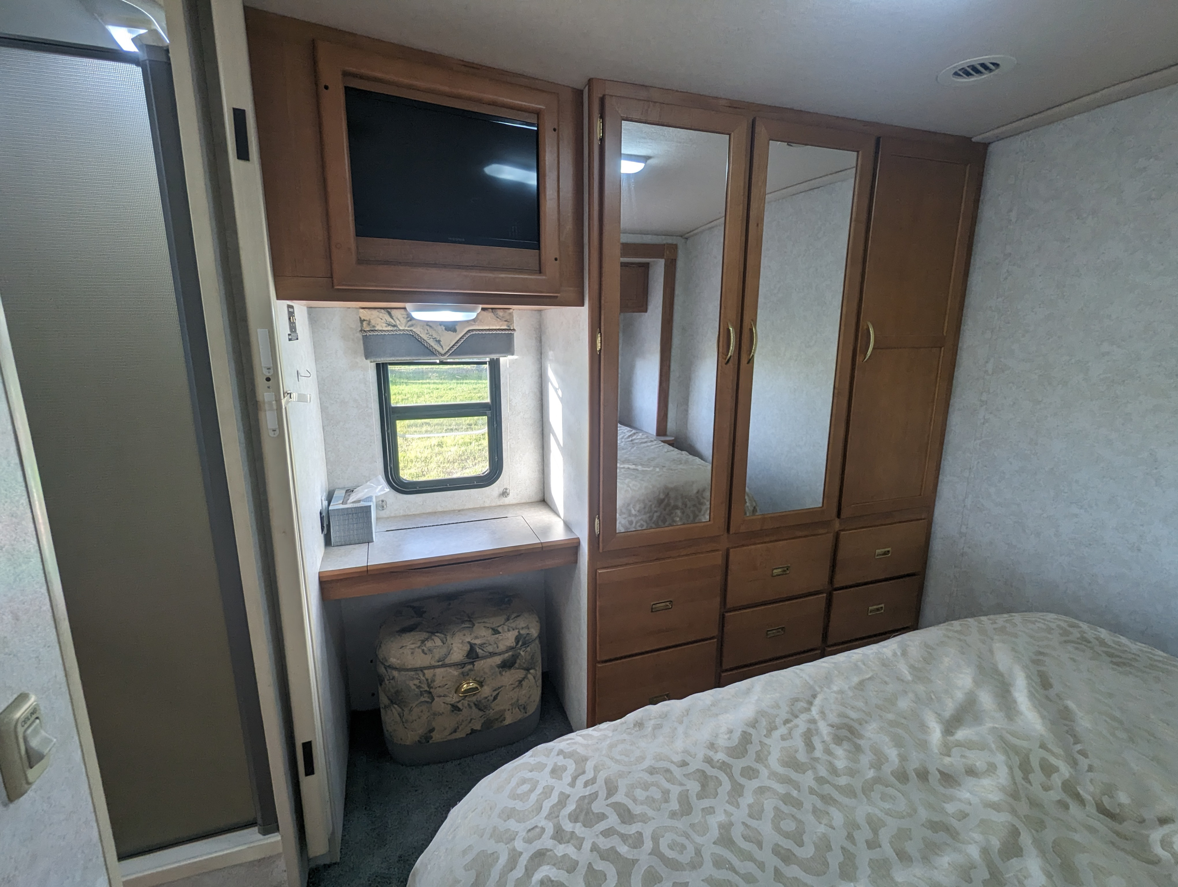 Inside an RV rental bedroom: cozy bed, built-in TV, desk with window view, and ample mirrored wardrobe storage.. Itasca Sunova 2006