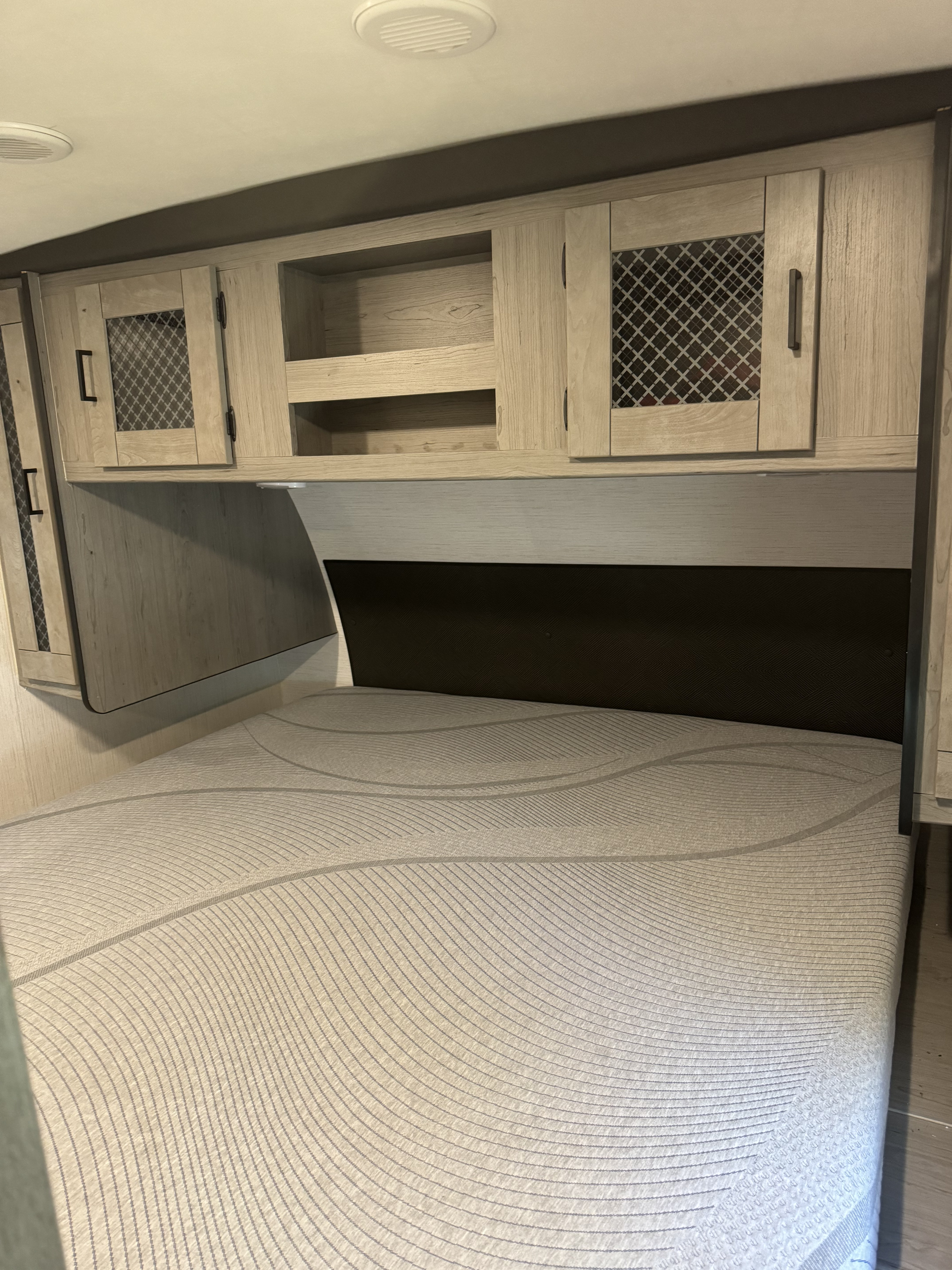 Experience RV rental comfort: a bedroom with a patterned mattress, wood-look overhead cabinets with mesh accents, and a dark headboard.. Heartland RVs Mallard 2022