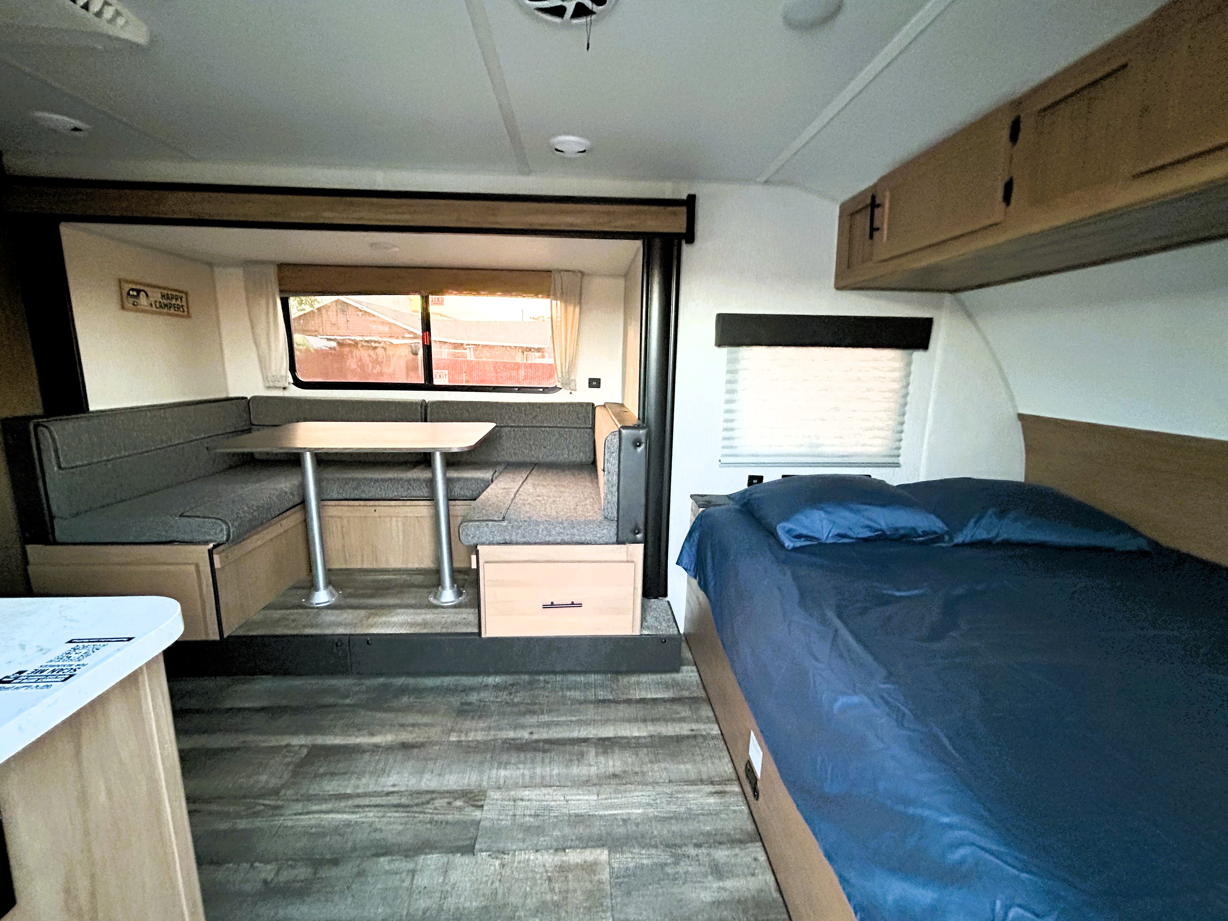 Modern rv rental interior: comfortable dinette, cozy bed with blue sheets, wood-look floor, and overhead cabinets.. Forest River Cherokee Wolf Pup 2023