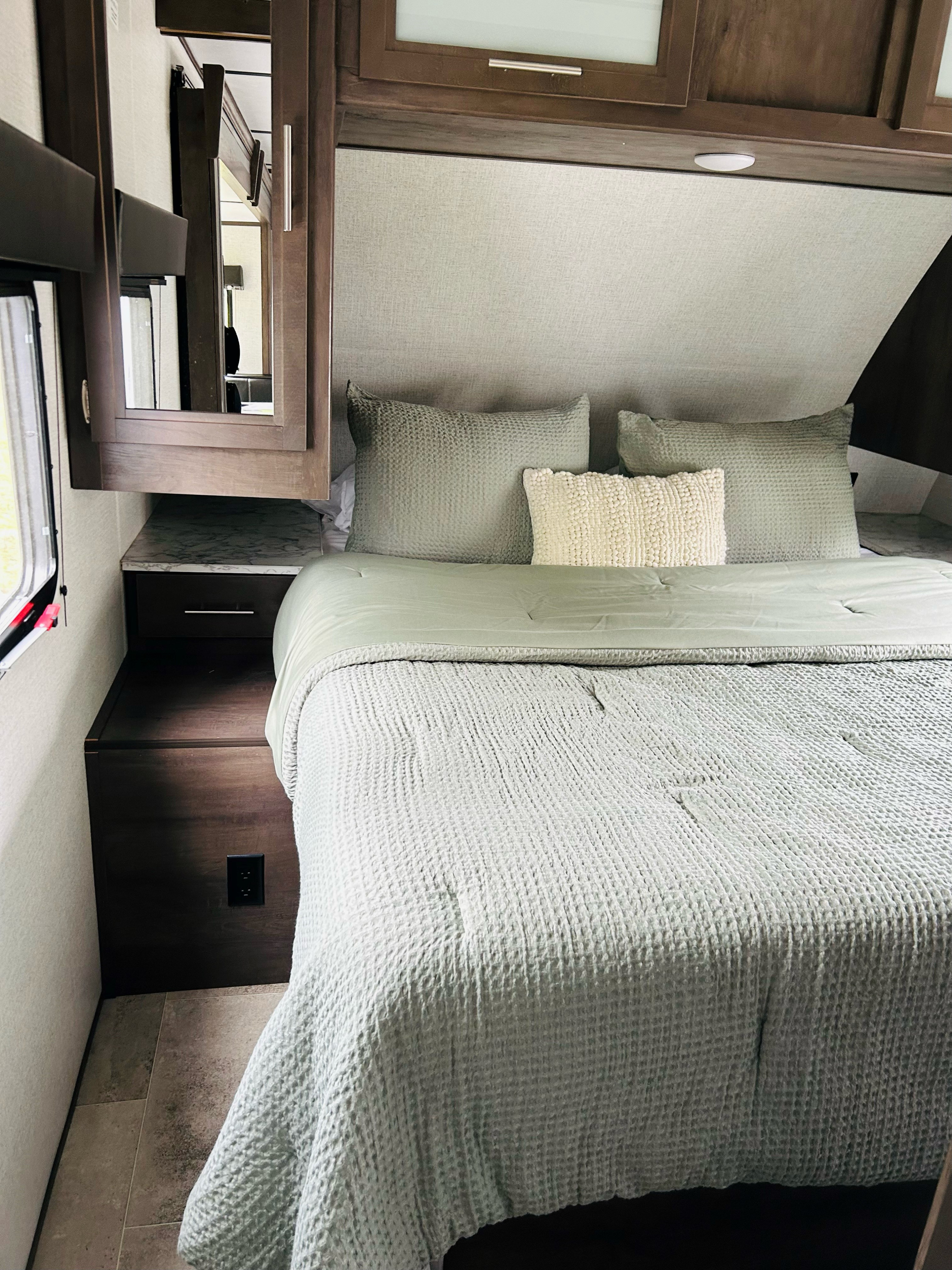 Inside a modern RV rental bedroom: a comfortable bed with sage green bedding, dark wood cabinets, and a nightstand.. Dutchmen Coleman Light 2021
