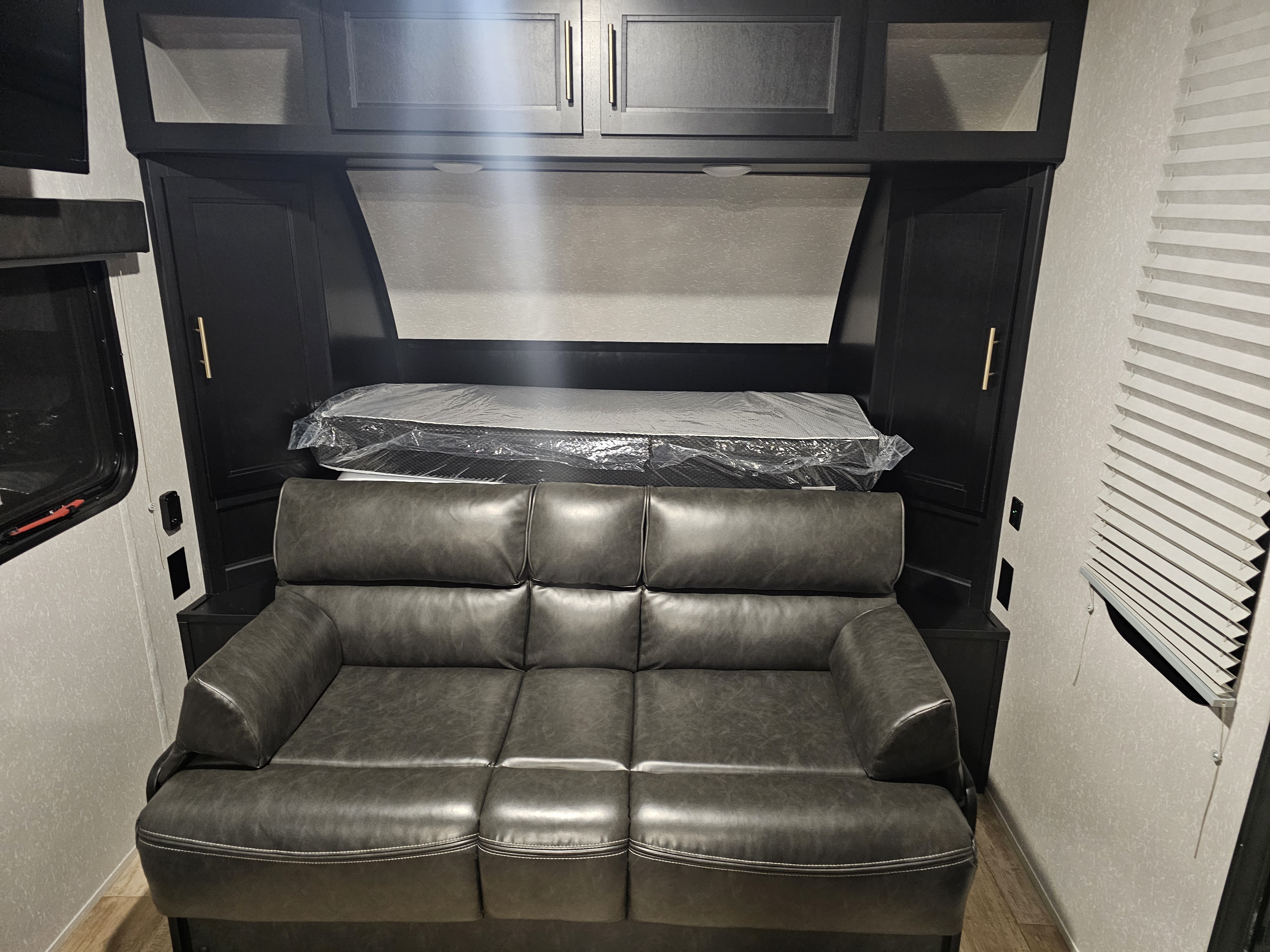 Inside an RV rental: a gray sofa below a plastic-wrapped mattress, dark cabinetry, and window blinds.. Forest River Cherokee Wolf Pup Black Label 2021
