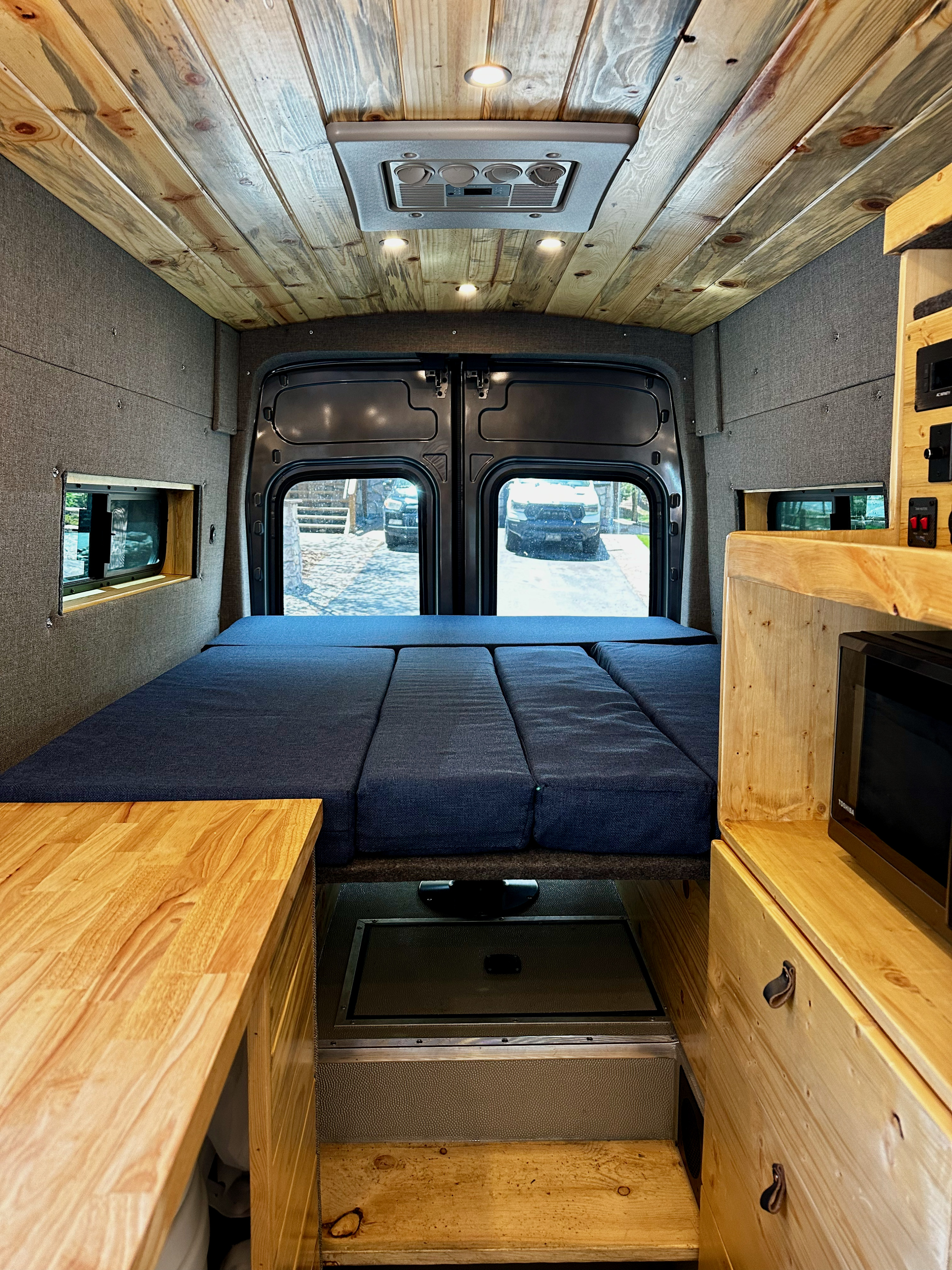 RV rental interior: A cozy camper van features a wood ceiling, plush blue bed, and integrated kitchen, ideal for van life travels.. Ford Transit 2023
