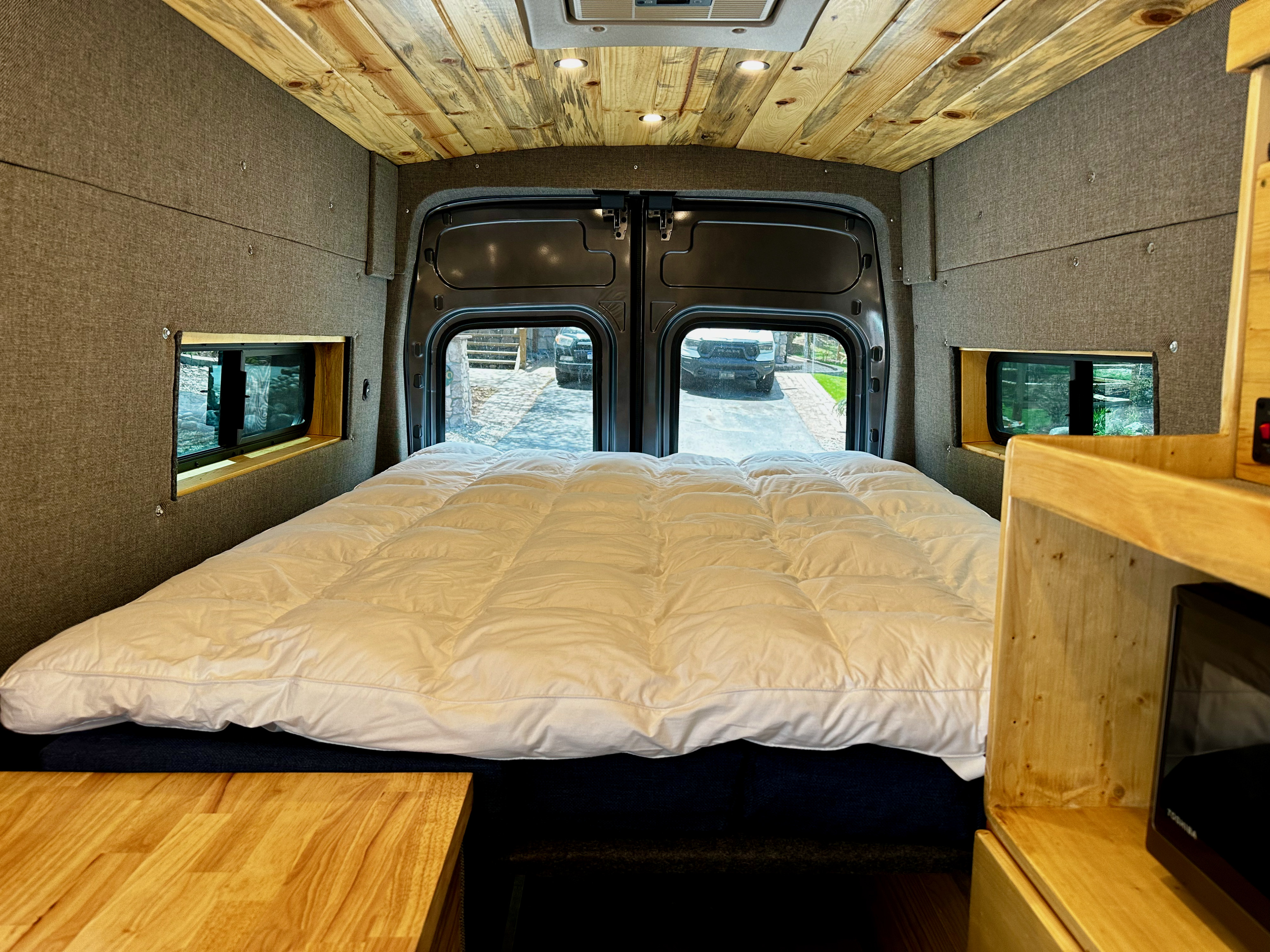 Cozy custom RV rental camper van interior. Features a comfortable bed with white comforter, wooden ceiling, upholstered walls, and rear windows.. Ford Transit 2023