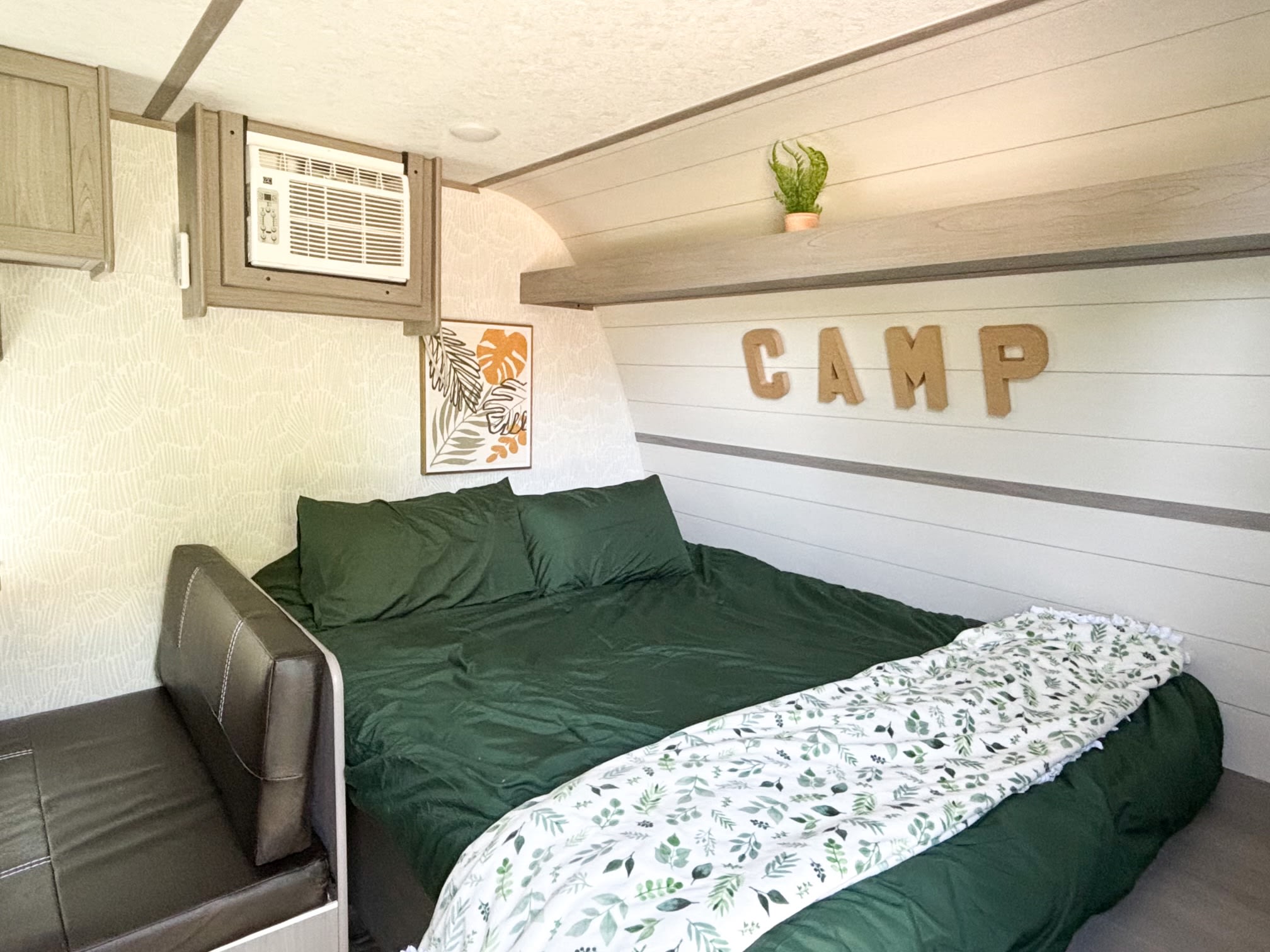 Cozy RV rental bedroom boasts green bedding, a leafy throw, and CAMP decor. Features include AC & bench seating.. Dutchmen Coleman 17B 2024