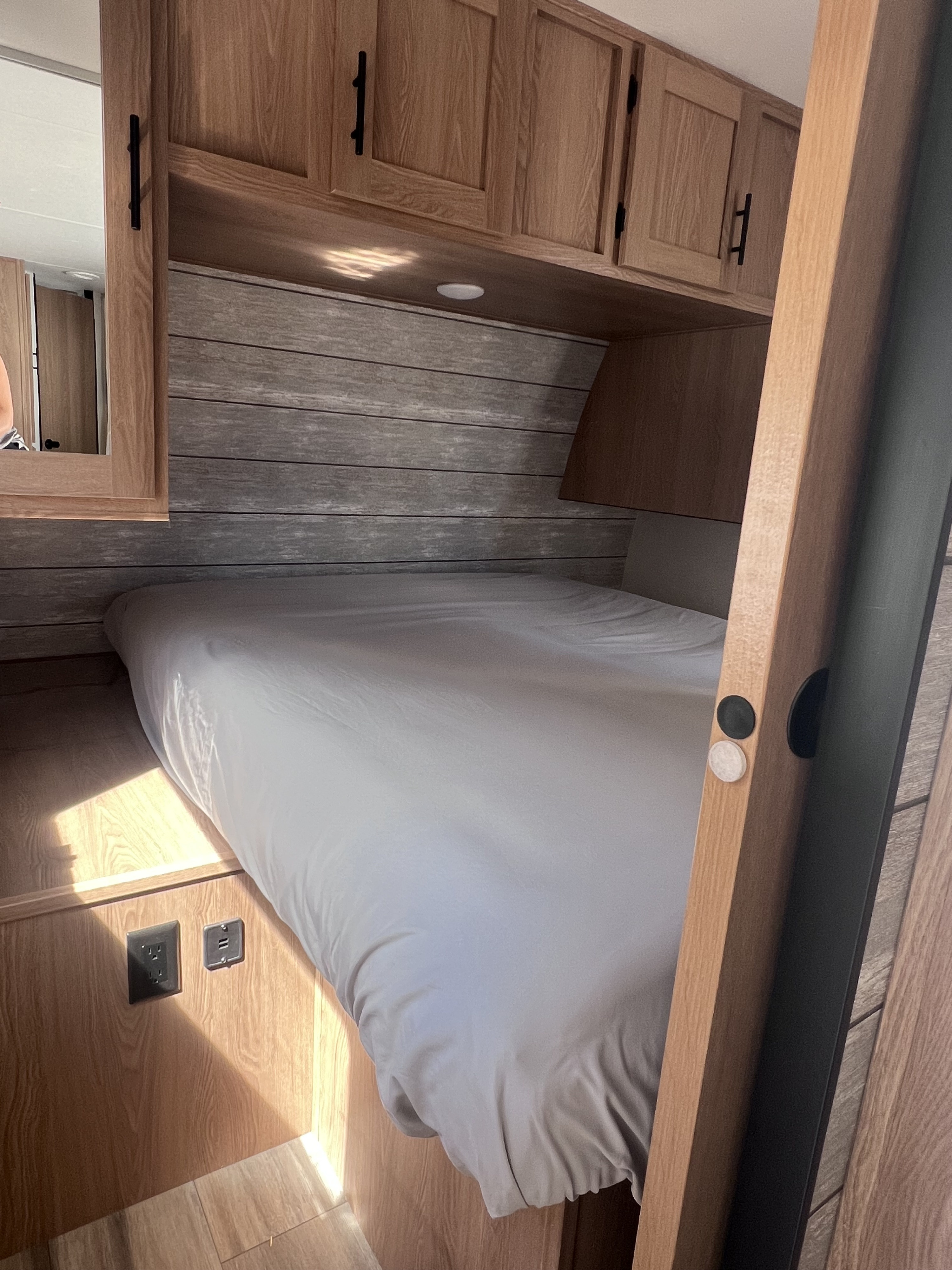 Inside an RV rental: a compact bedroom with a bed, wood-paneled walls, overhead cabinets, and convenient power outlets.. Prime Time Avenger 24 BHS 2023