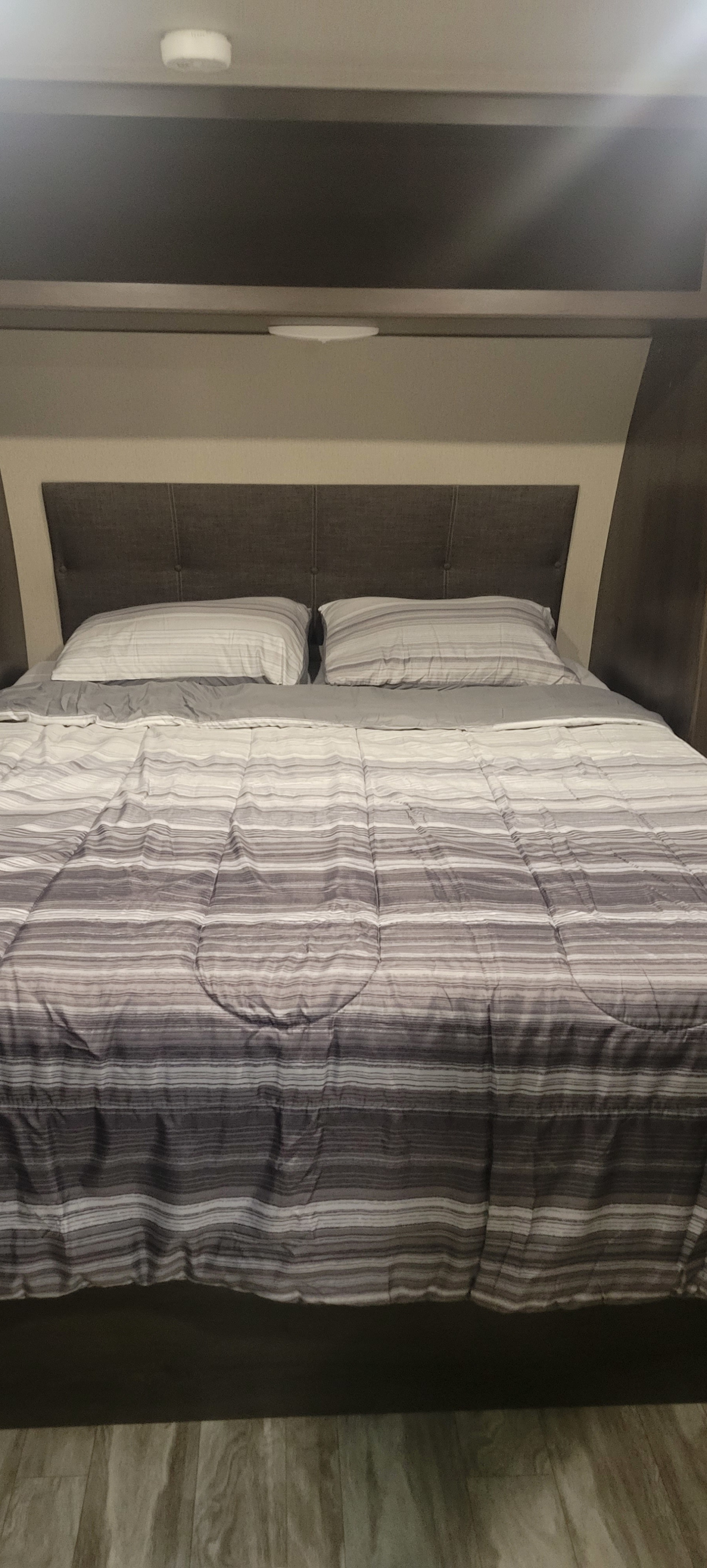 Cozy bed with striped bedding and a dark headboard in an RV rental's compact bedroom, featuring overhead lights and wood flooring.. Grand Design Transcend 2021