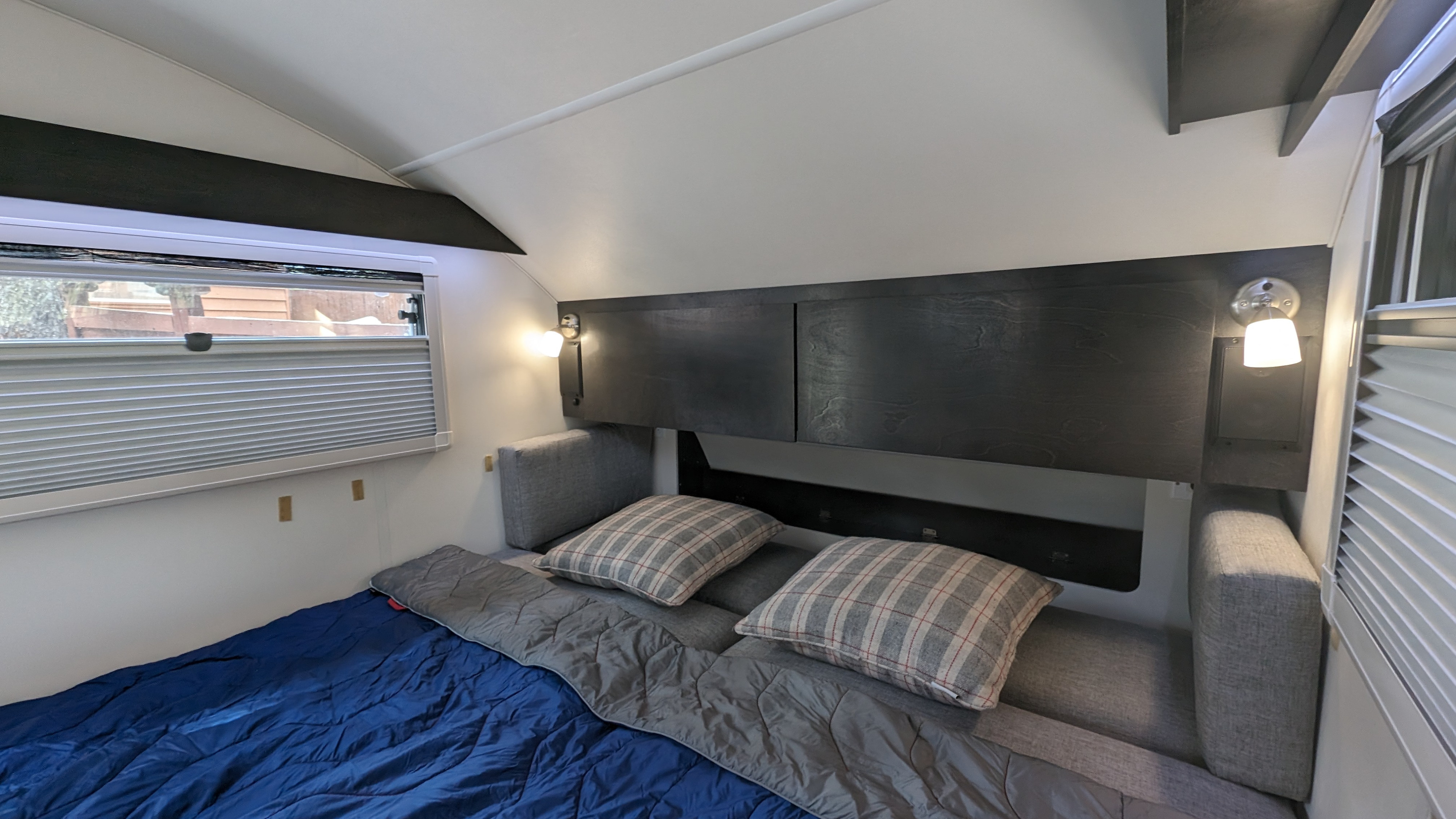 Comfortable RV rental interior featuring a neatly made bed with pillows, alongside a window with blinds and built-in lighting.. nuCamp T@G 2023