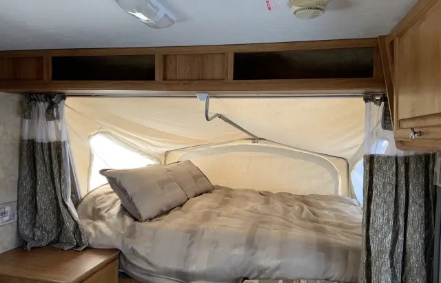 Interior of an RV rental showing a comfortable pop-out bed, light bedding, and patterned curtains.. Jayco Jayfeather 2007