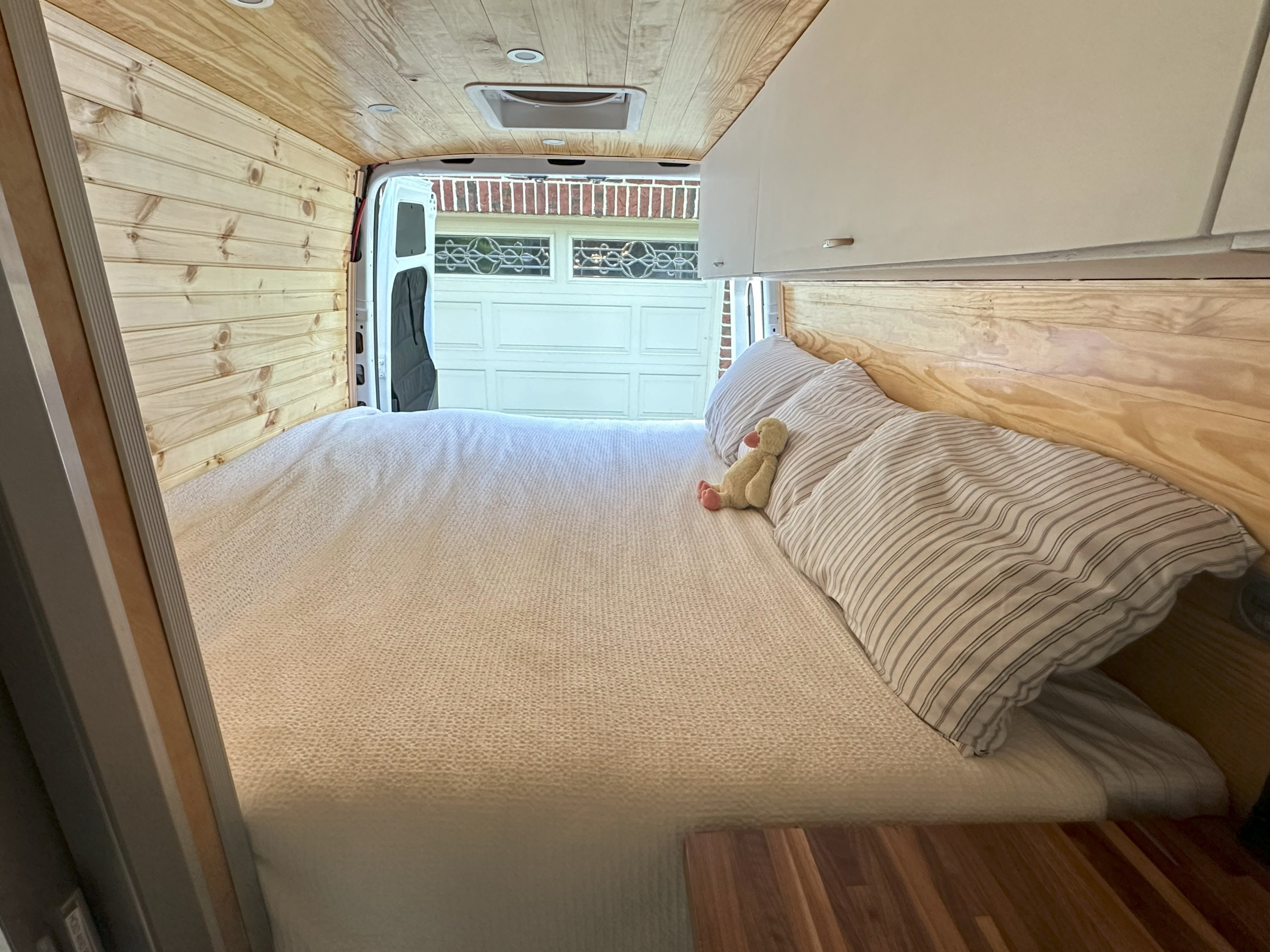 Cozy camper van interior for RV rental. Features a spacious bed, wood paneling, and overhead storage, open to a garage view.. Mercedes-Benz Sprinter 2021