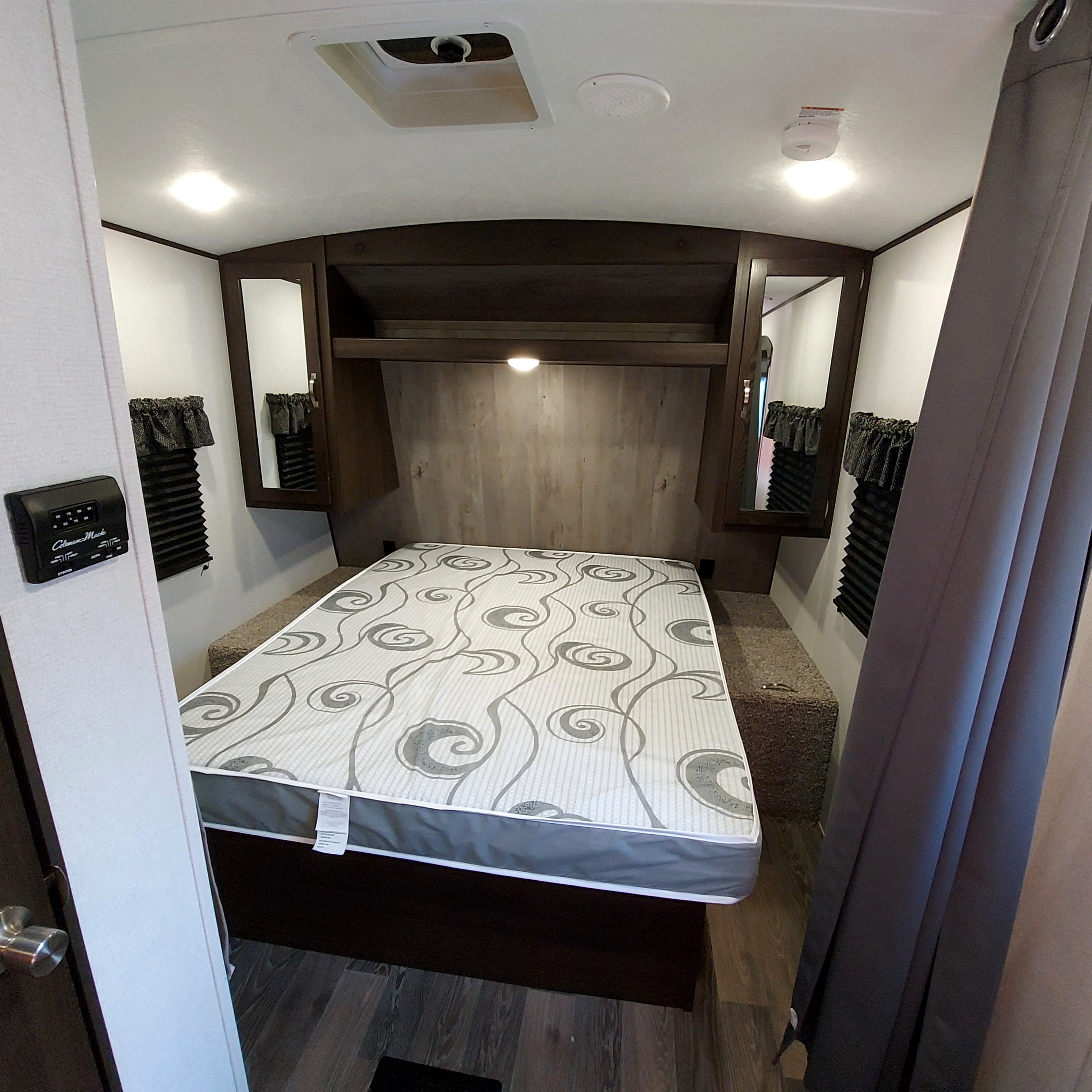 An RV rental bedroom with a patterned mattress, overhead storage, side mirrors, windows with blinds, and ceiling fixtures.. Keystone Springdale 2021