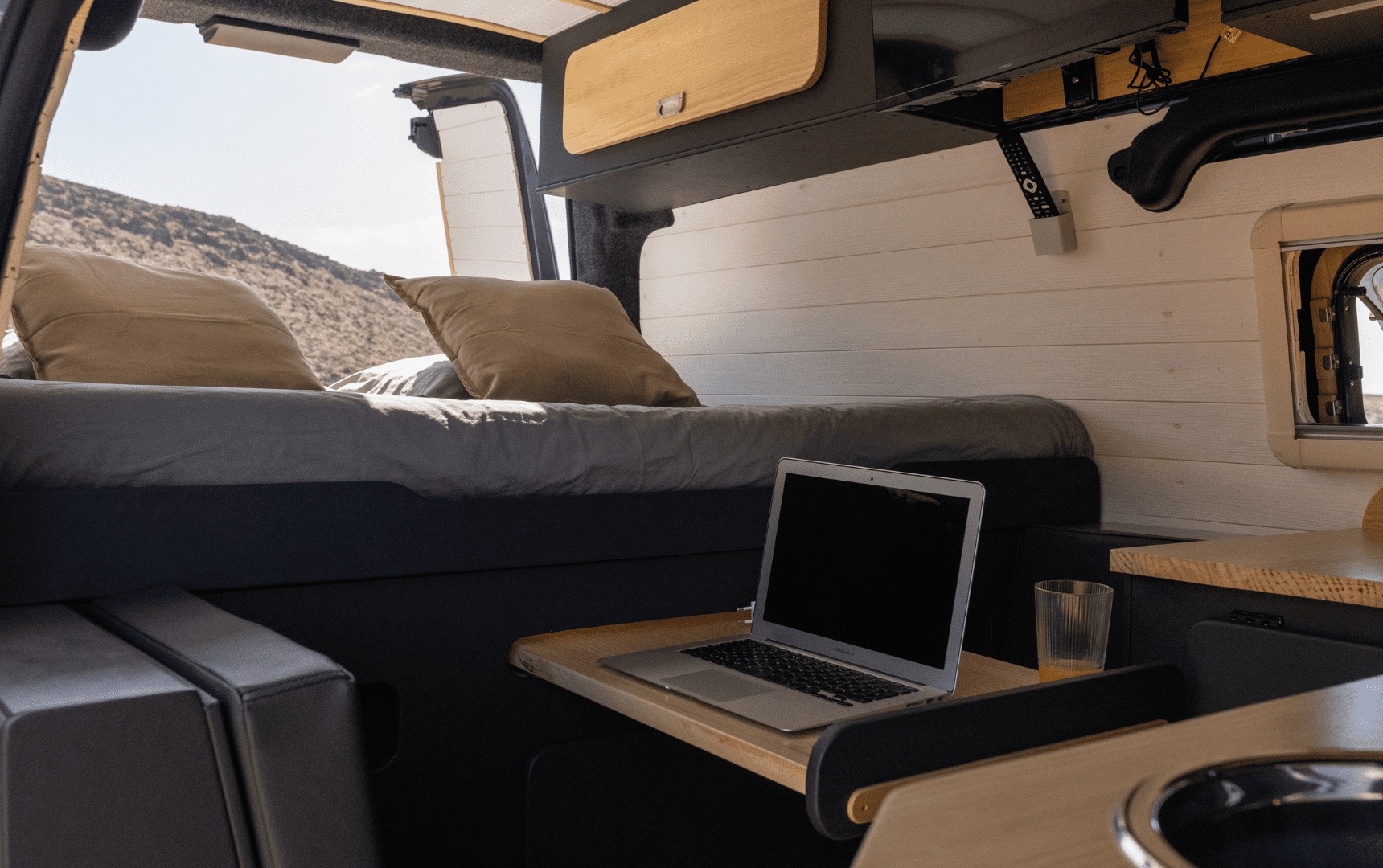 Inside a modern RV rental, featuring a comfortable bed, mobile office setup with a laptop, and a compact kitchen.. Ford Transit Custom 2023