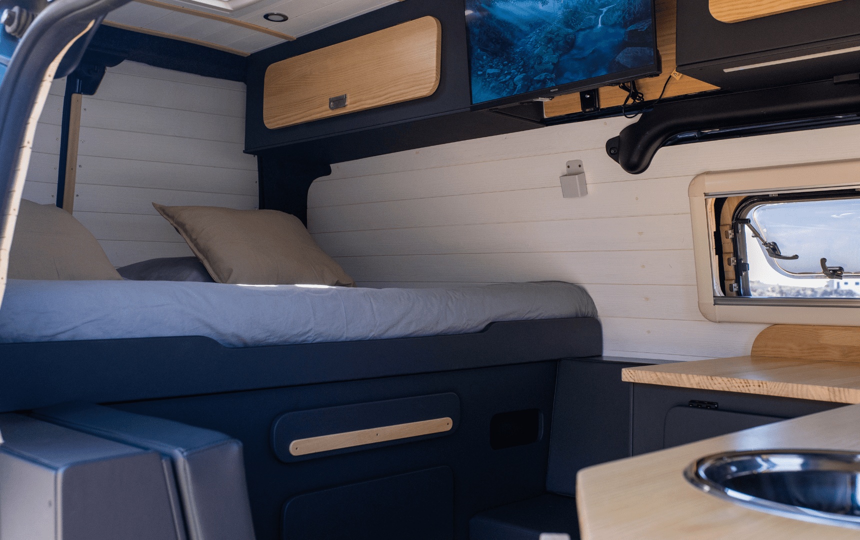 Comfortable RV rental interior: a bed with pillows, wall-mounted TV, wood cabinets, and a kitchen sink are visible.. Ford Transit Custom 2023