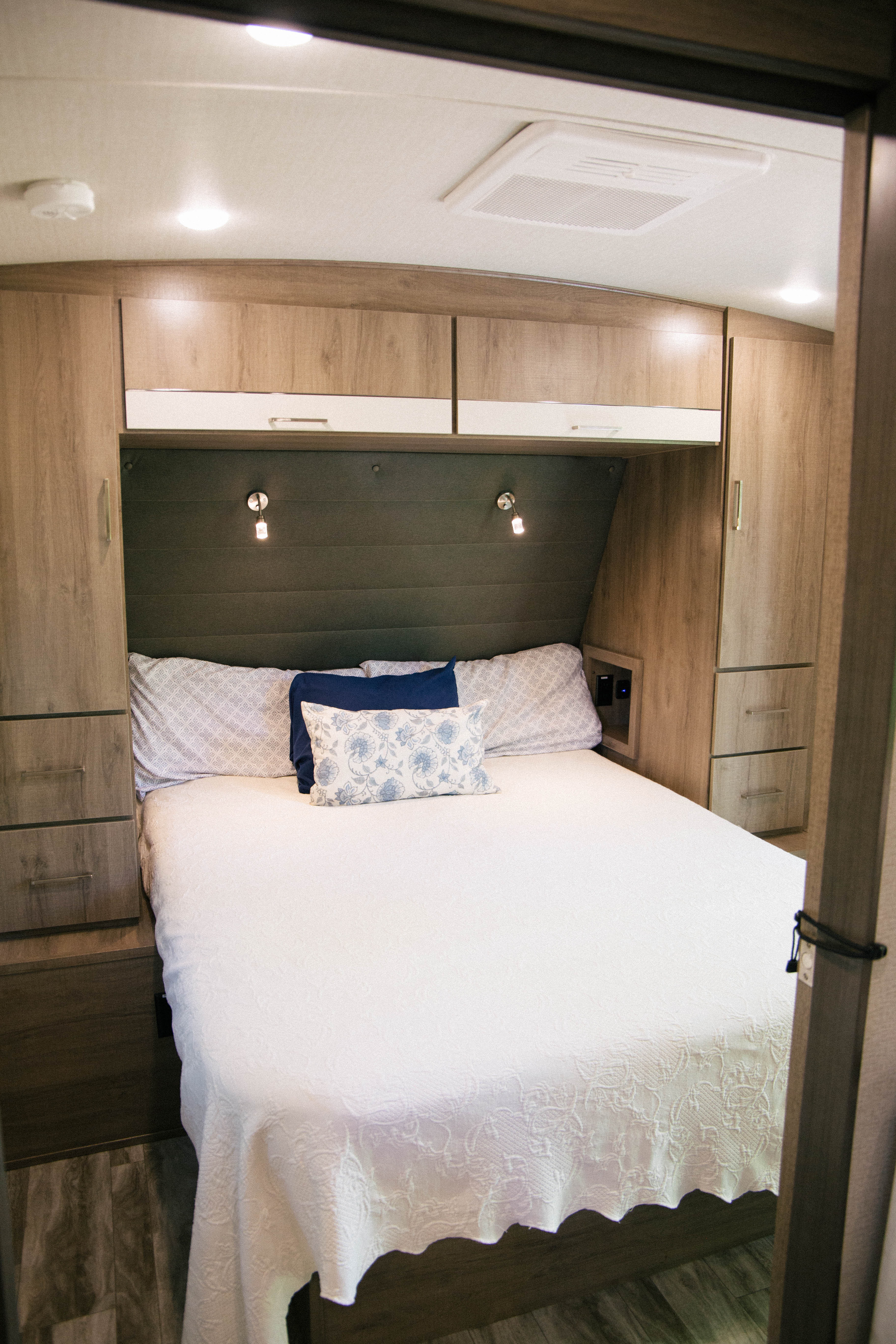 An RV rental bedroom with a neatly made bed, built-in wood cabinets, and functional lighting.. Grand Design Imagine 2023