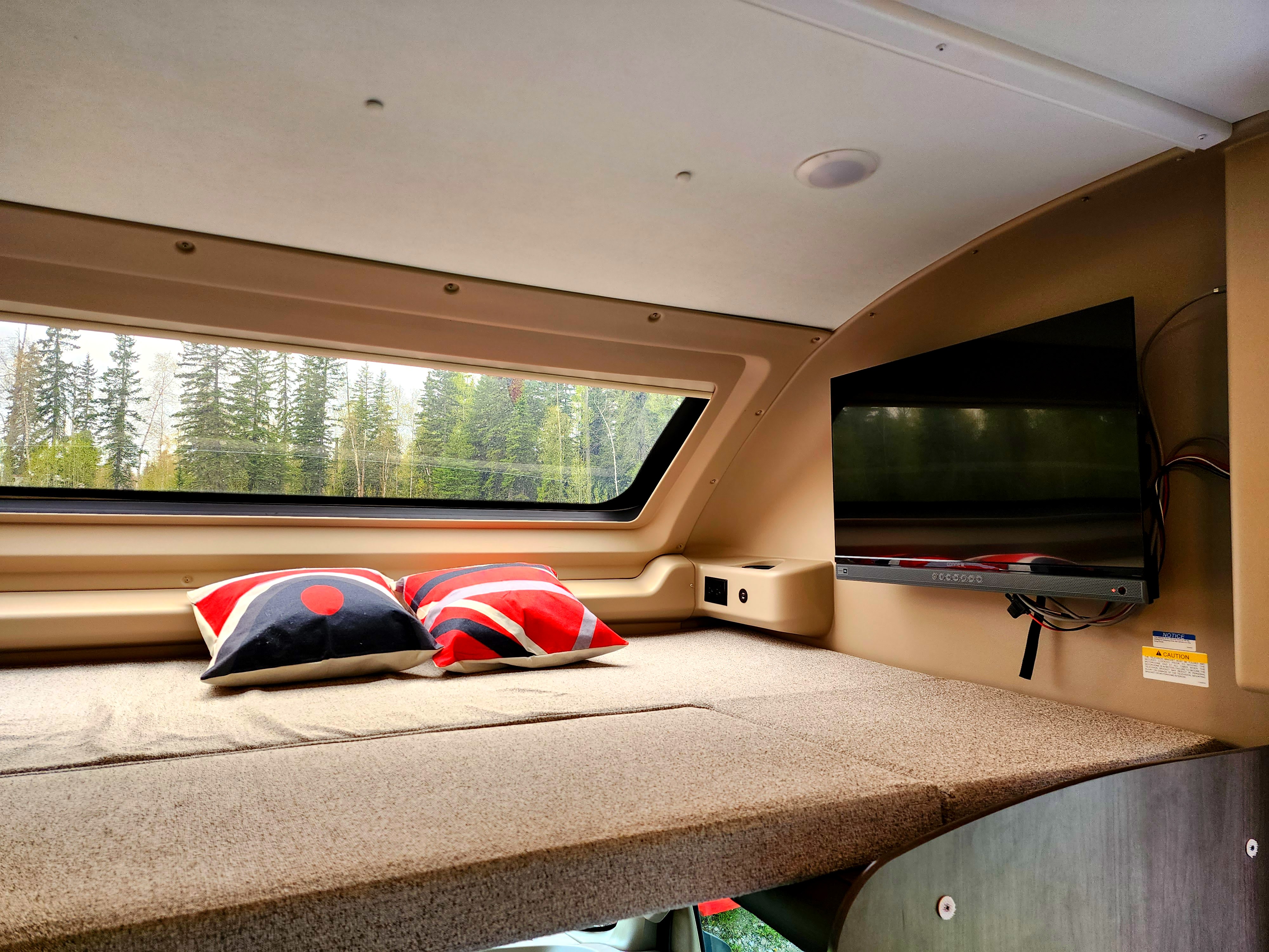 Inside a cozy RV rental: A comfortable sleeping area with a large window offering forest views, a TV, and charging ports.. Jayco Entegra Odyssey 2024