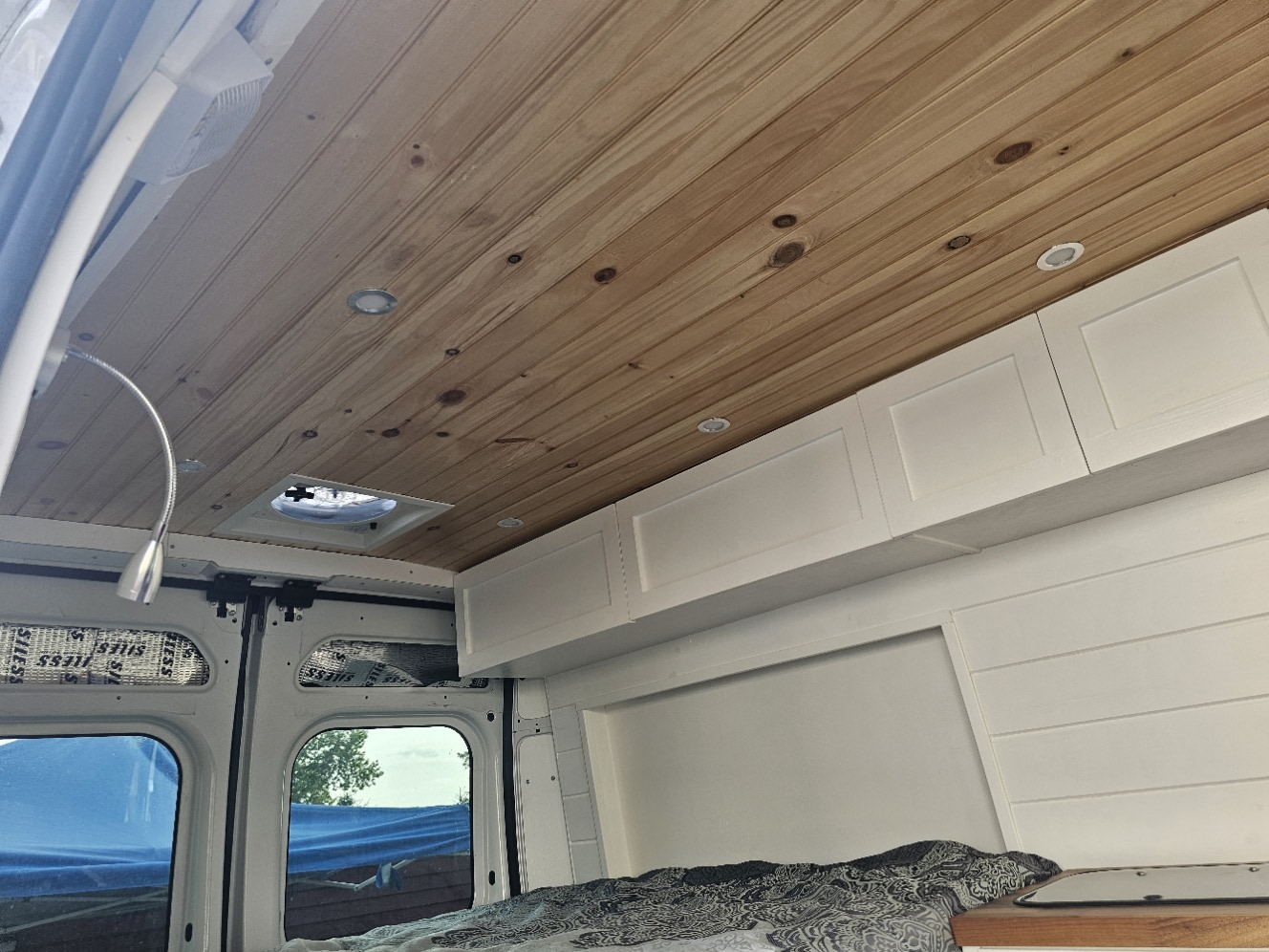 RV rental: Bright, modern camper van interior with wood plank ceiling, white overhead cabinets, flexible lamp, and cozy bed.. RAM Promaster High Top Built in spring 2024 2016