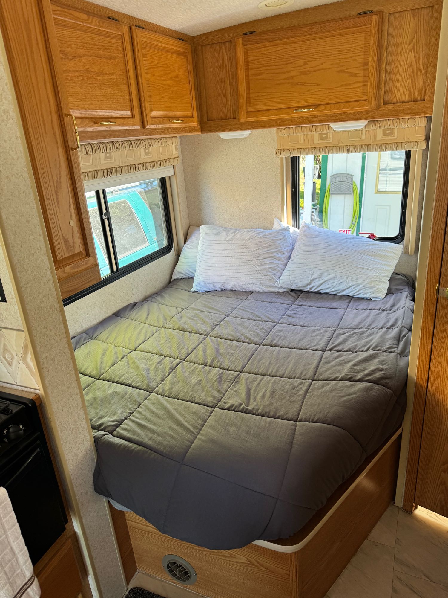 Inside an RV rental: a comfortable bed with grey comforter, overhead wood cabinets, and two windows for scenic views.. Winnebago Minnie 2004