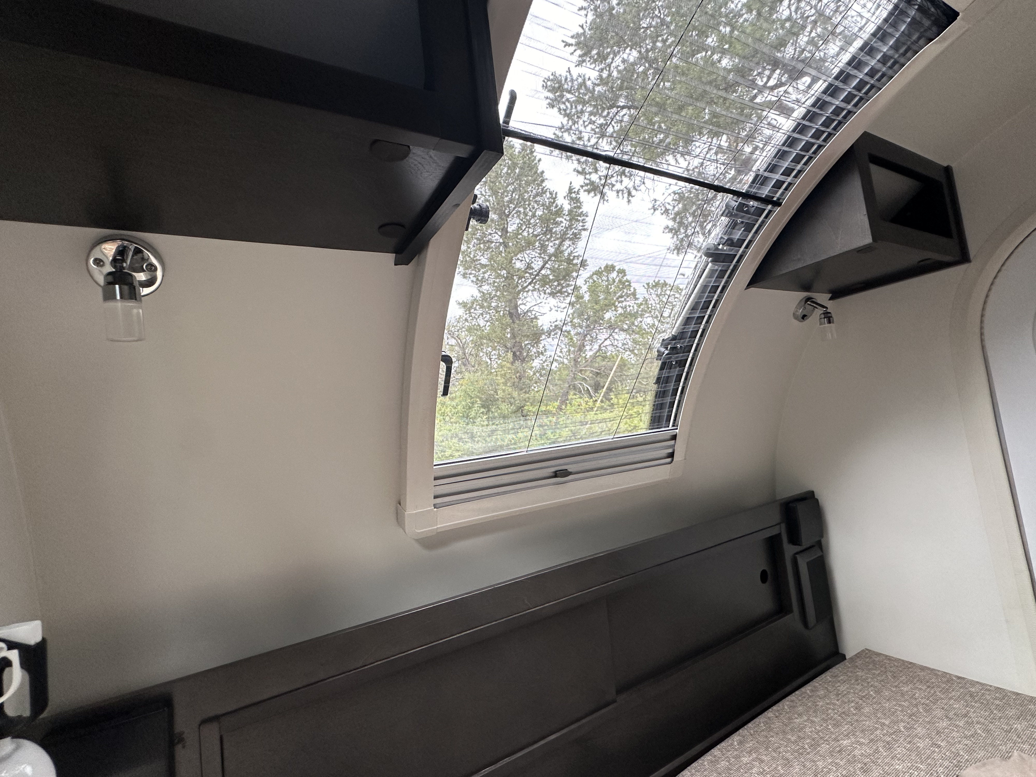 Cozy RV rental interior with a comfortable bed, overhead cabinet, and a large window overlooking trees, featuring a pull-down screen.. nuCamp T@G 2021
