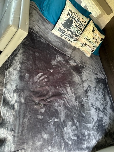 Inside a comfy RV rental: a bed with camping-themed pillows & a textured gray blanket showing handprints.. Keystone RV Bullet Crossfire 2021