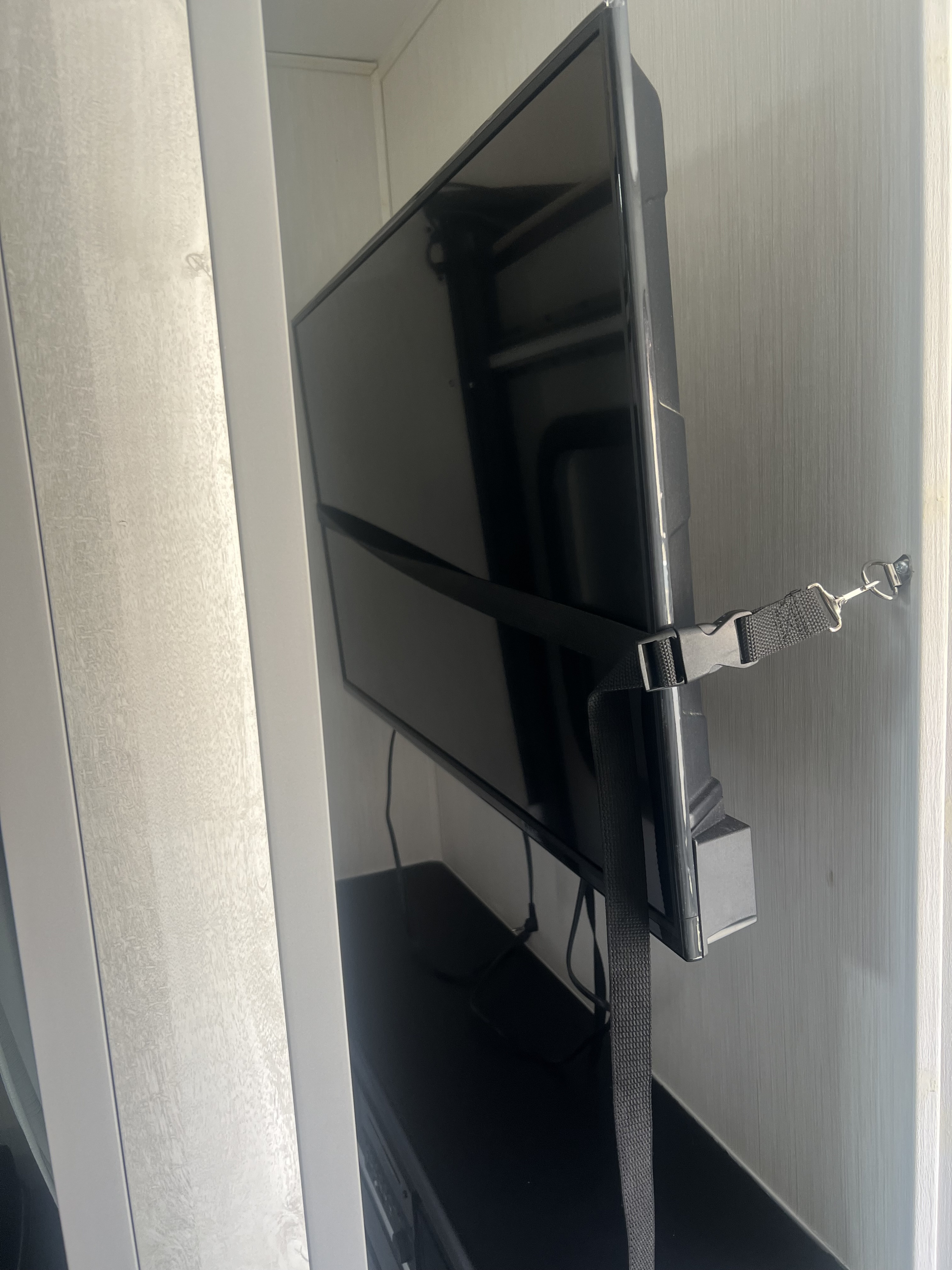 In an **RV rental**, a flat-screen TV is secured with a black strap and metal clip to the textured wall, preventing movement during travel.. Forest River Vibe 2021