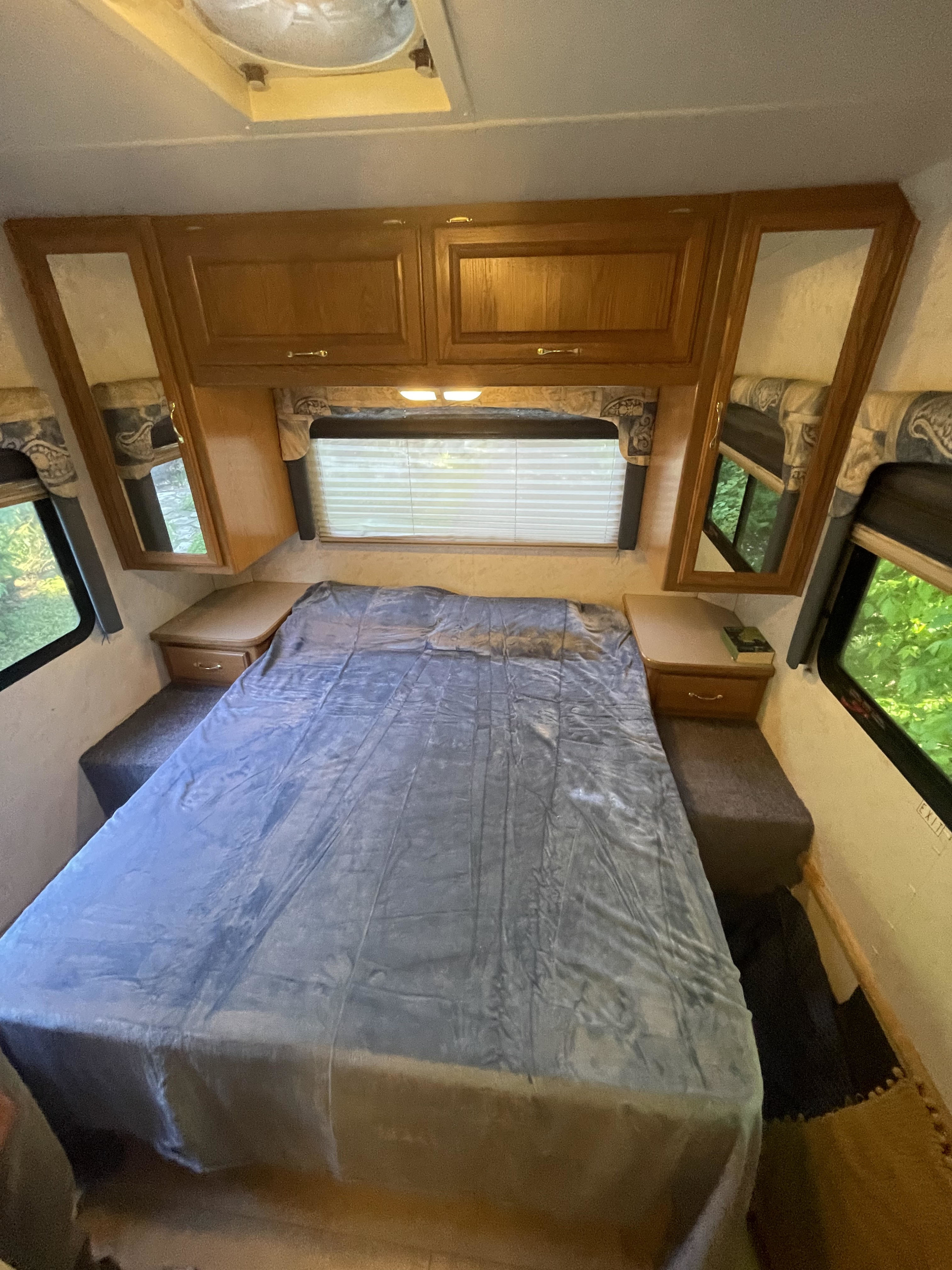 Inside an RV rental bedroom: blue blanket on bed, wooden overhead cabinets, and windows with outdoor views.. Four Winds 5000 2002
