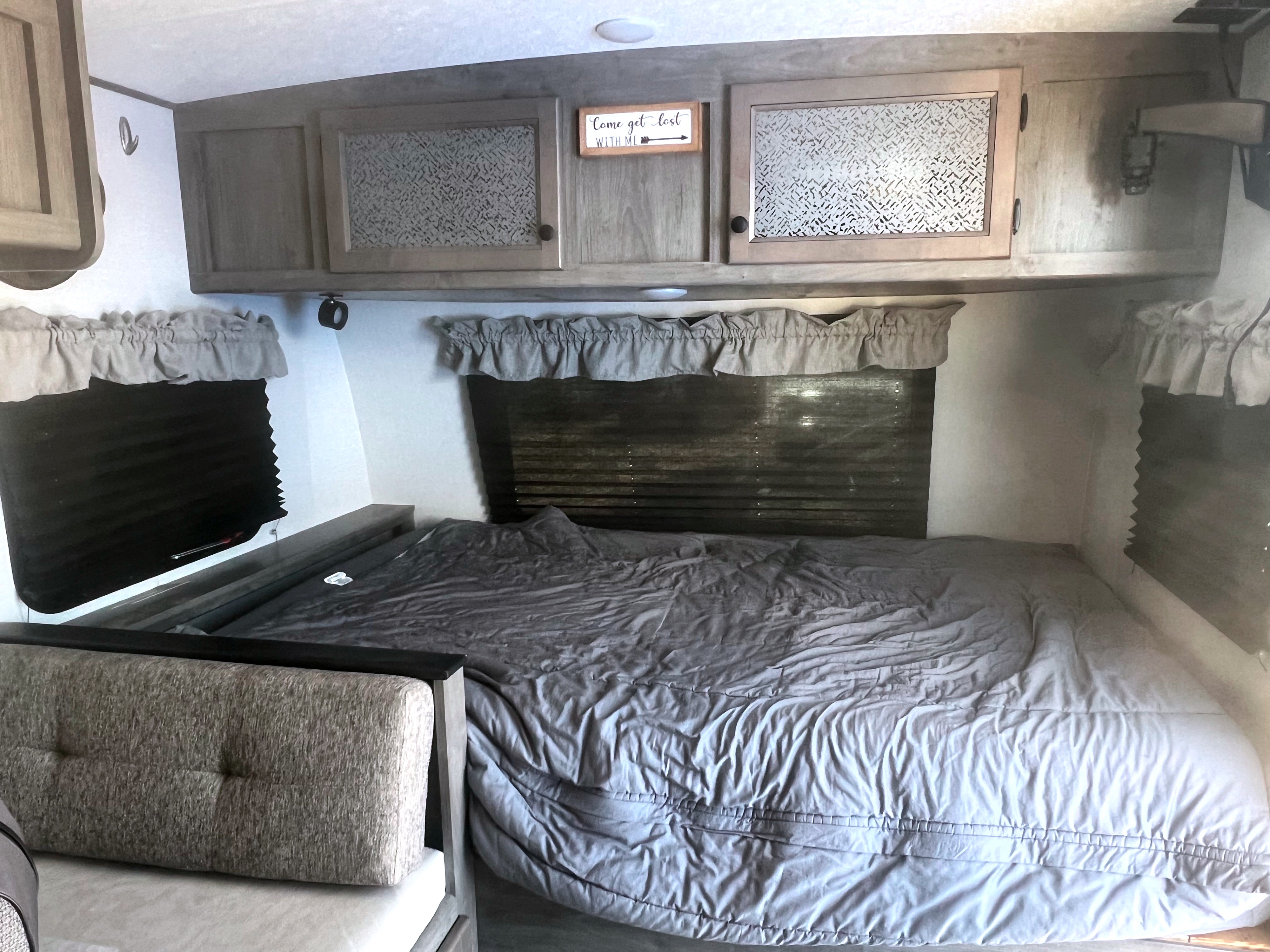 An RV rental bedroom with cozy gray bedding, overhead cabinets, a "Come get lost" sign, and window shades.. Coachmen Apex Nano 2021