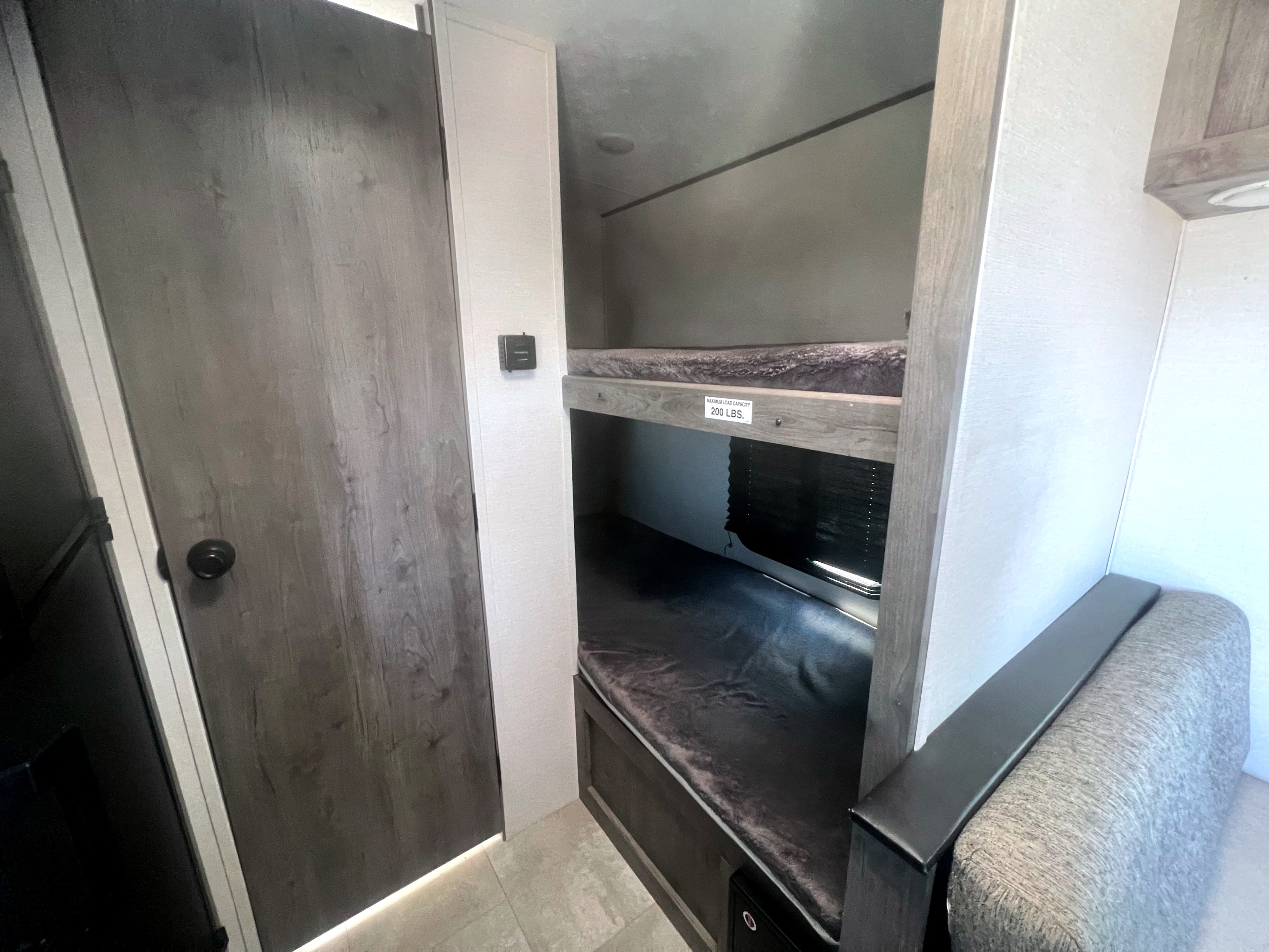 Inside an RV rental: two cozy bunk beds with a 200 lb. capacity, a wood-look door, and an adjacent seating area.. Coachmen Apex Nano 2021