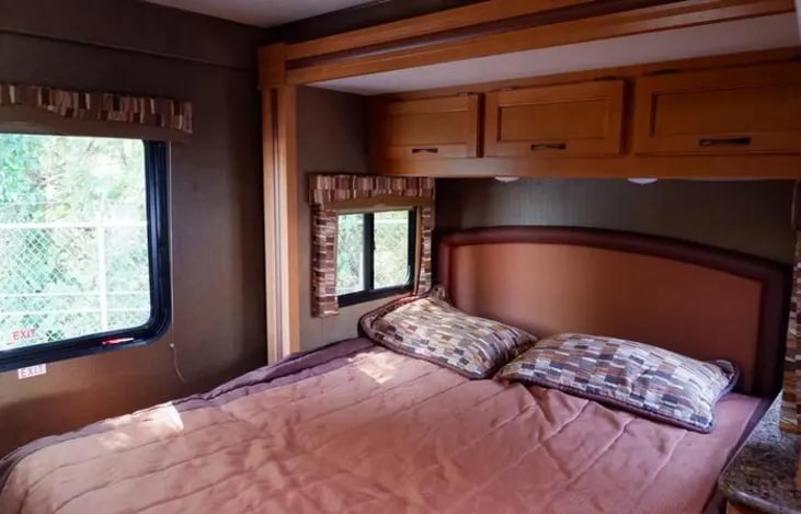 Inside an RV rental: a compact bedroom with a bed, patterned pillows, wood cabinets, and a window. Ready for your next adventure.. Thor Chateau 2017