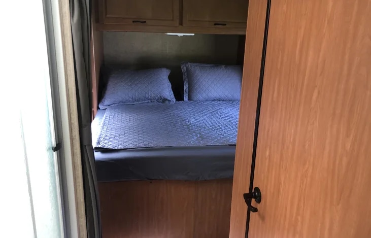 Cozy RV rental bedroom featuring a blue quilted bed, pillows, and wooden overhead cabinets. Ideal for travel.. Thor Majestic 2016