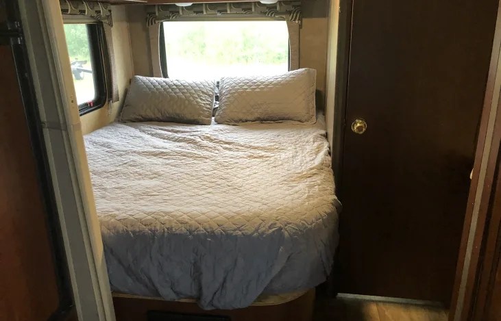 Cozy RV rental bedroom with quilted bedding, a large window revealing greenery, and a wooden door.. Forest River Sunseeker 2016