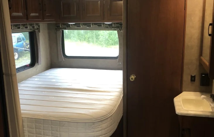 RV rental interior: bedroom with a large mattress, windows overlooking a truck and trees, overhead cabinets, and a small sink.. Forest River Sunseeker 2016