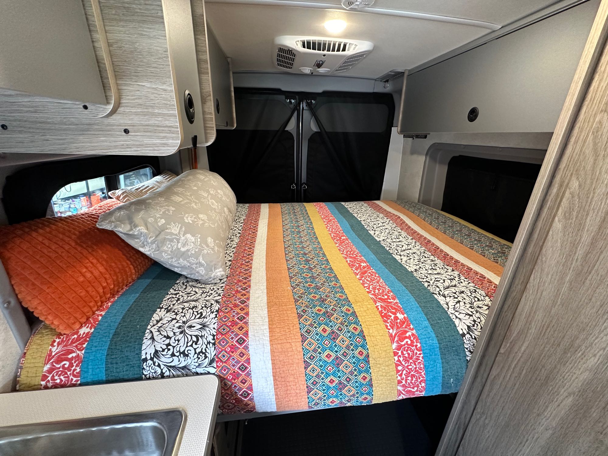 RV rental interior: A vibrant, striped quilt covers the bed. Overhead cabinets, a ceiling AC unit, and a sink are visible.. Winnebago Solis 2023